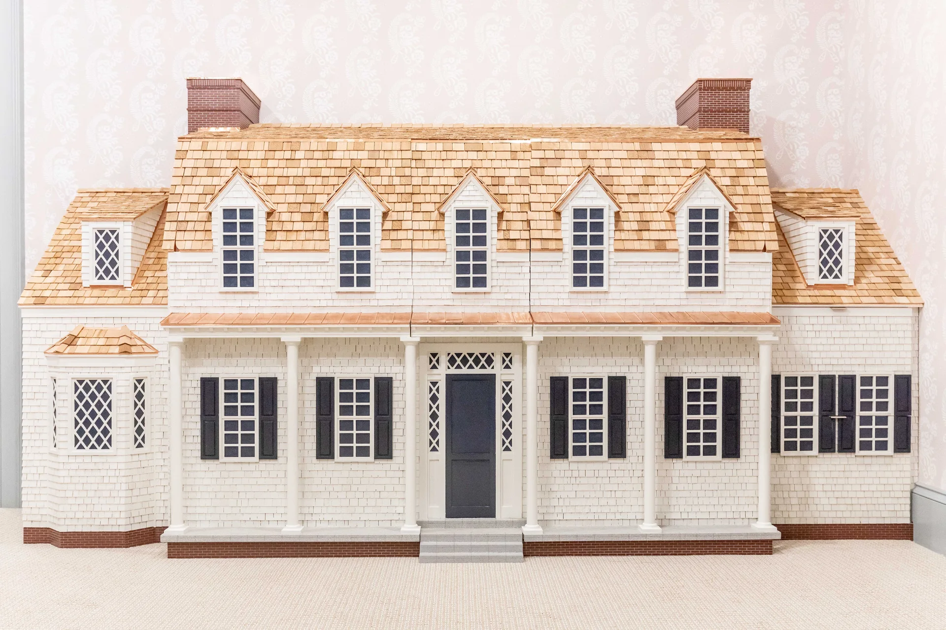 A miniature colonial style dollhouse features a symmetrical white facade, dark navy shutters, wood shingle roof, and dual brick chimneys.