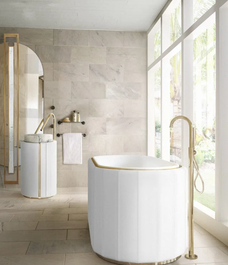 A modern cream bathroom features a channel-tufted freestanding bathtub, a matching pedestal sink, gold hardware, and large stone wall tiles.