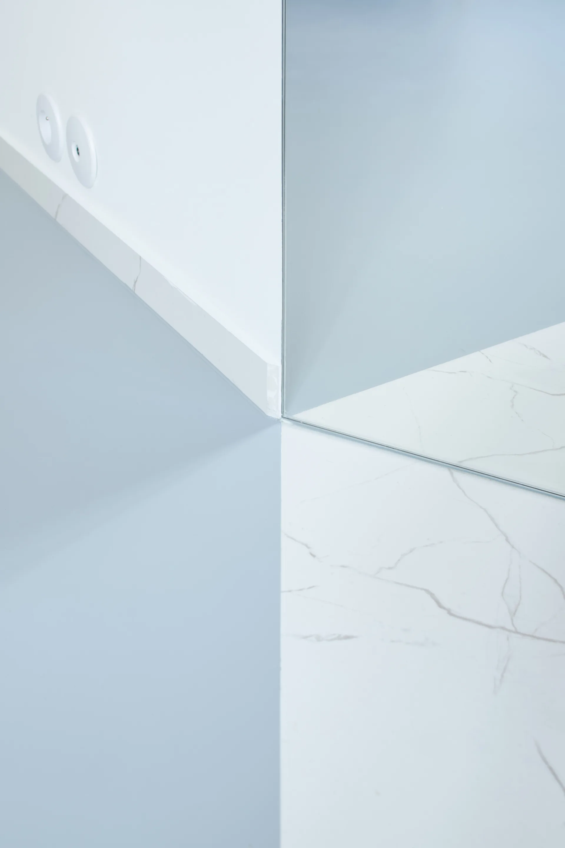 Close-up of an architectural corner showing a white wall, a floor-to-ceiling mirror, and a transition between blue and white marble flooring.