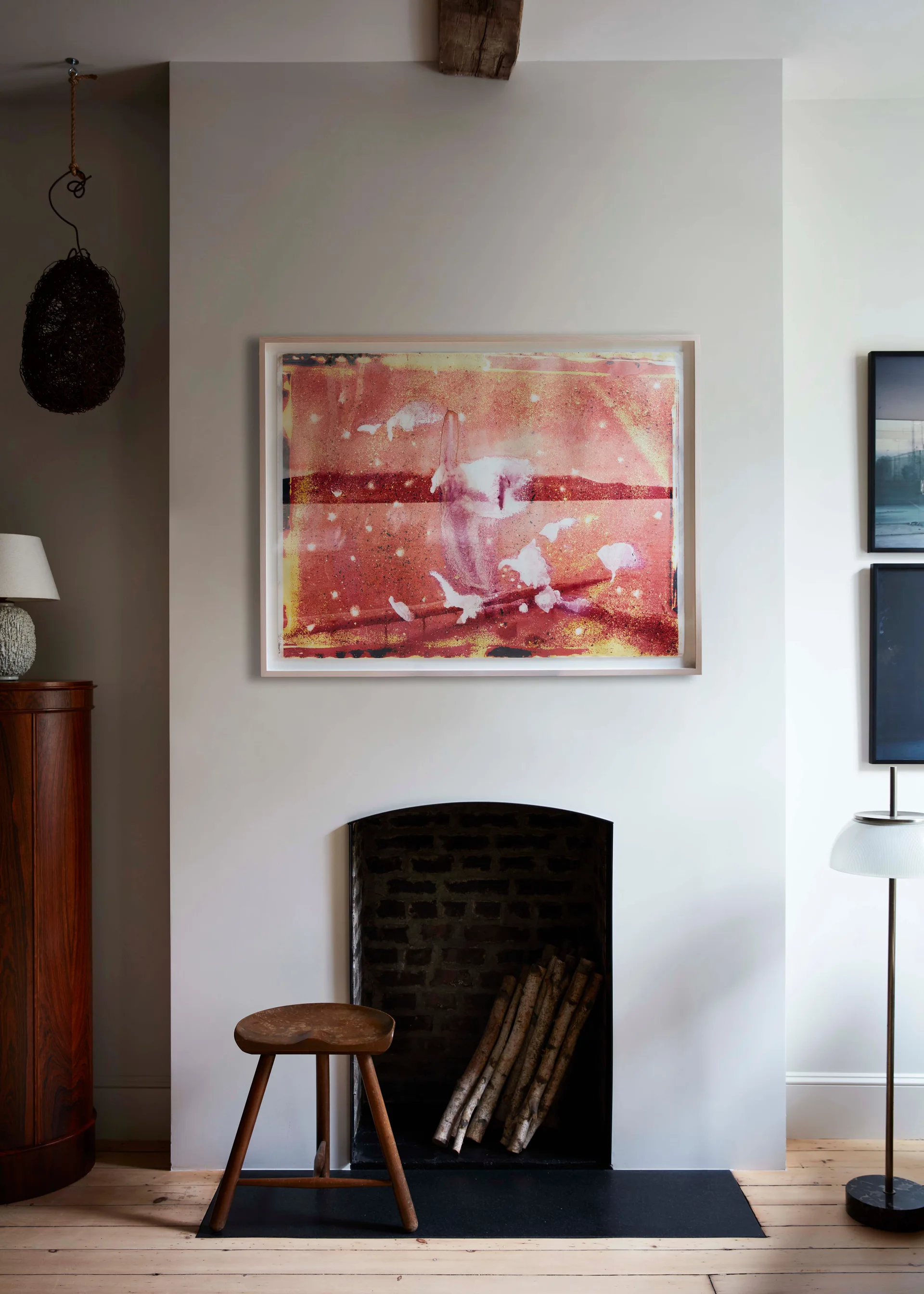 A minimalist grey fireplace wall features an arched brick firebox with birch logs, a wooden stool, and a large red abstract framed painting above.
