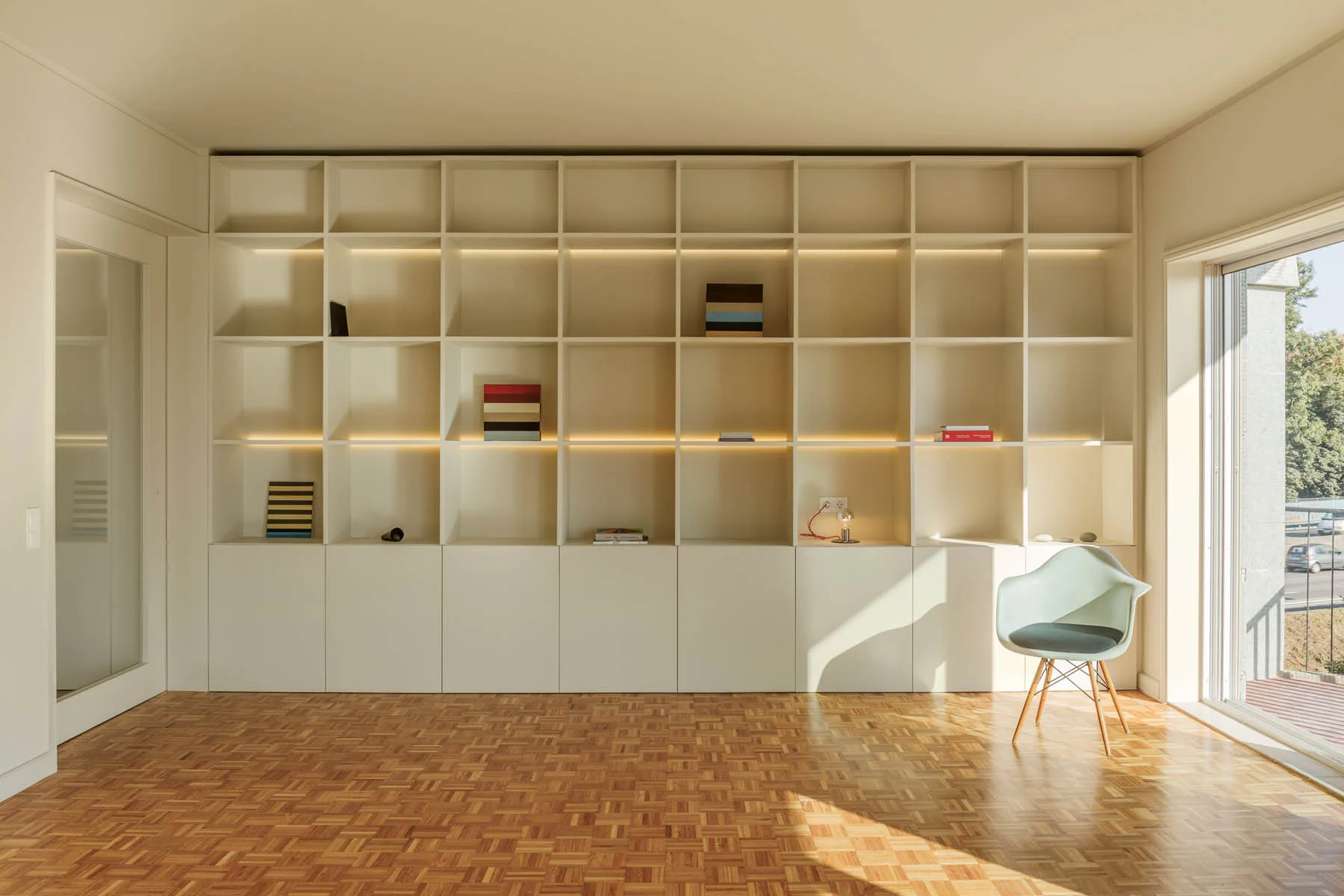 A large, built-in cream shelving unit with integrated lighting stands against a wall in a room with a patterned wood parquet floor and a modern chair.