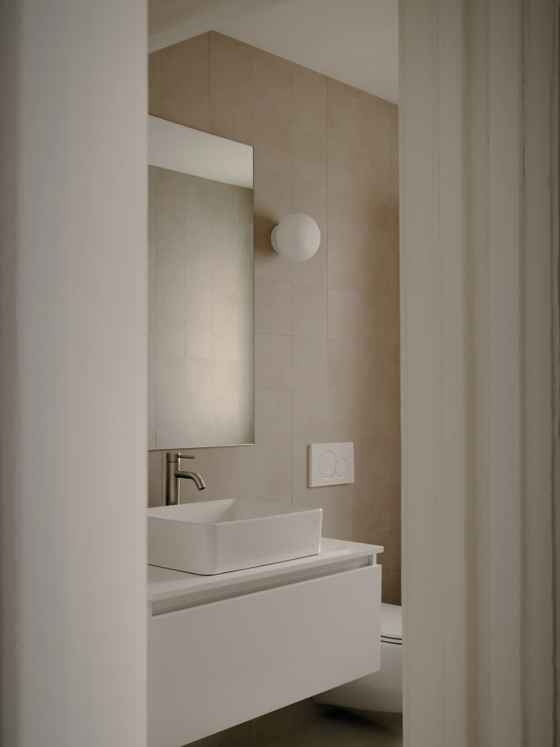 A minimalist bathroom features a white vessel sink on a floating vanity set against a wall of matte beige square tiles.