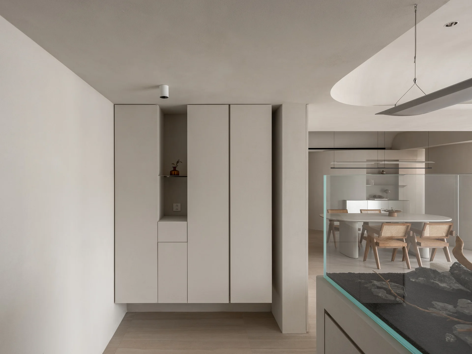 A minimalist beige dining space features floor-to-ceiling built-in cabinets, a wood dining table with rattan chairs, and a glass partition.