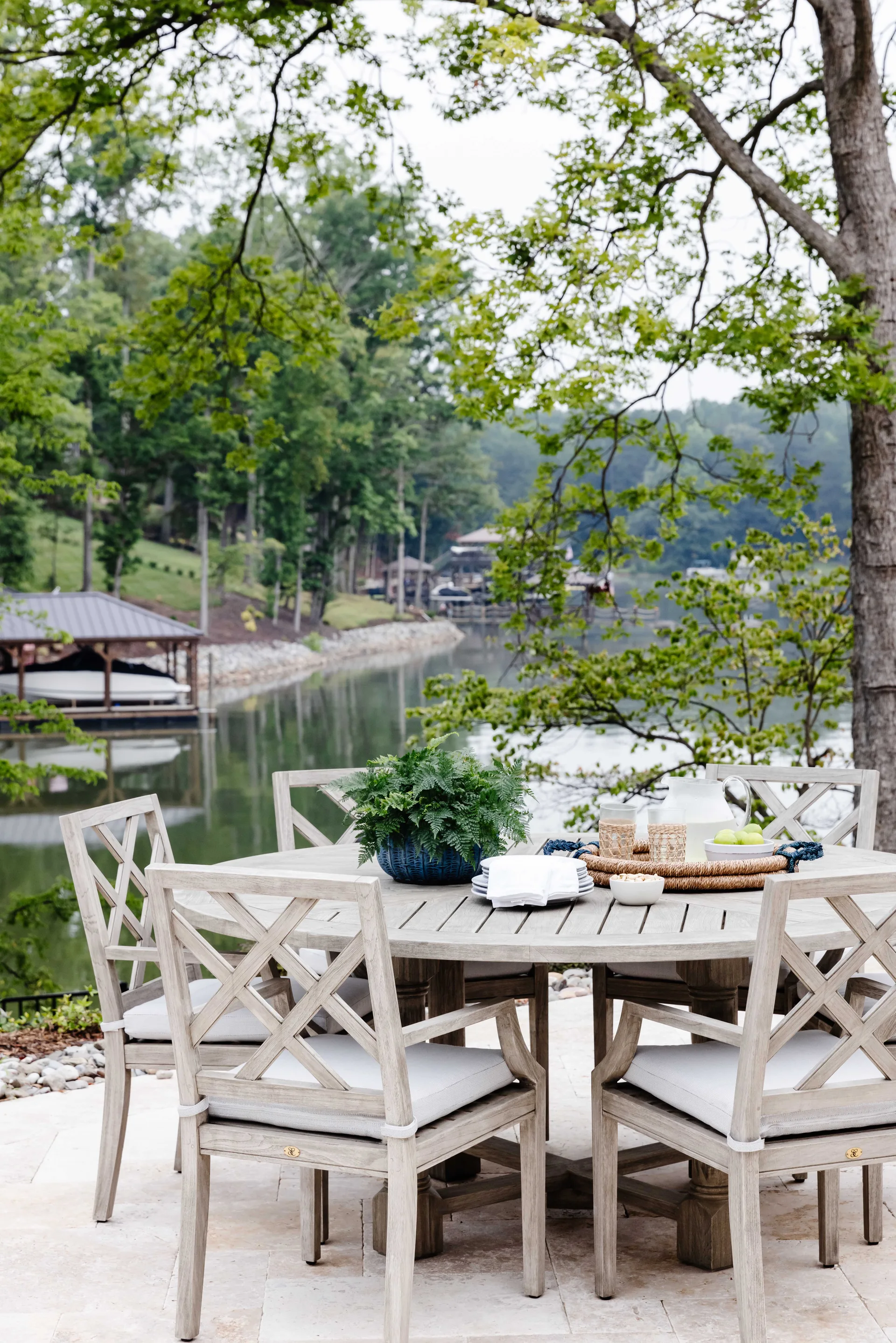 A circular weathered wood table and matching x-back chairs sit on a light stone patio overlooking a lake surrounded by green trees.