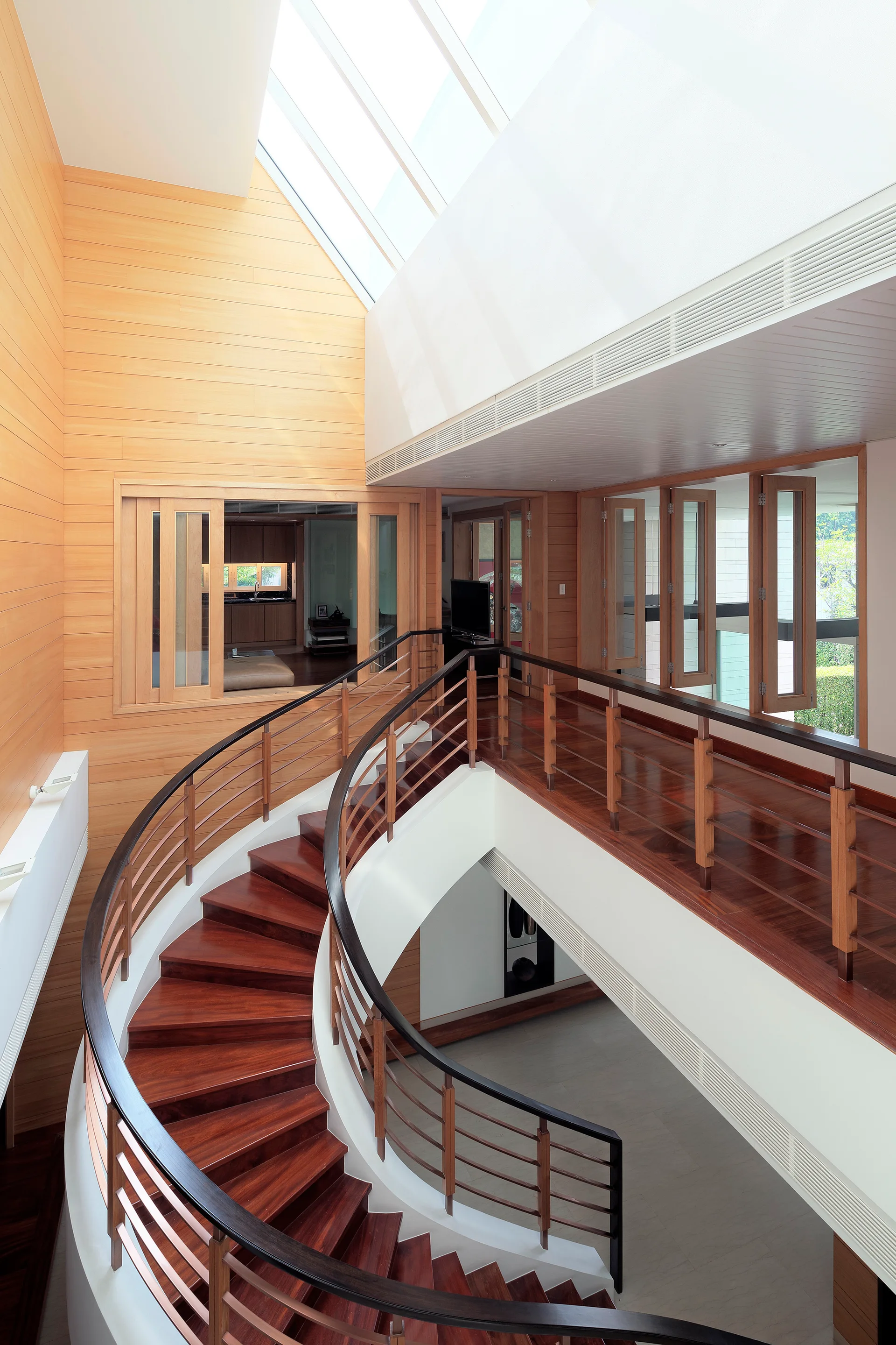 A curved dark wood staircase descends through a bright atrium with horizontal light wood wall paneling and a large angled skylight.