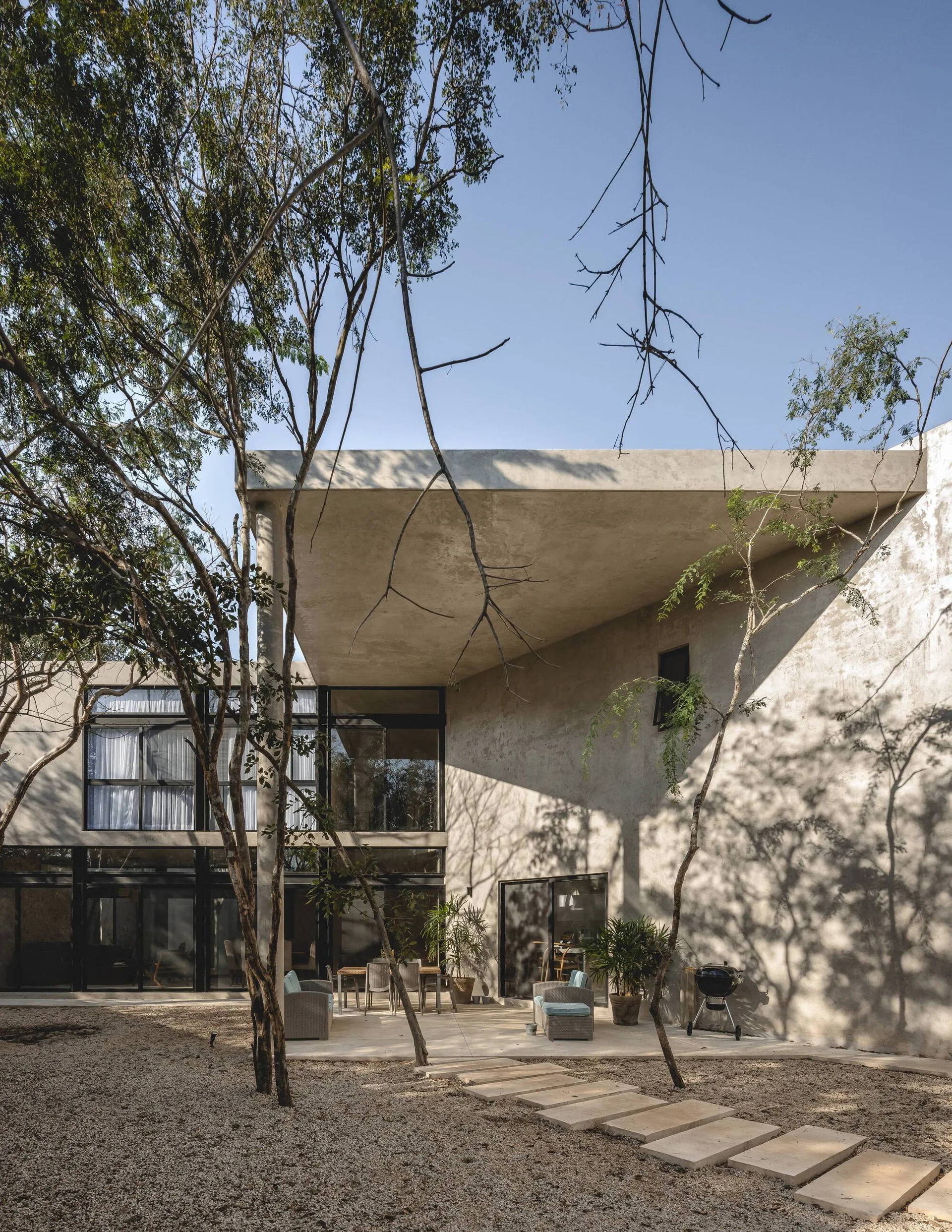 A modern residence with a massive concrete overhang shading an outdoor patio with dining and lounge furniture set on stone pavers.