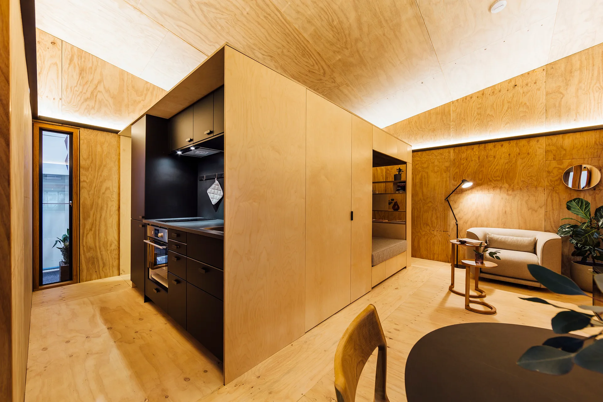 A compact interior with plywood walls and ceiling featuring a matte black kitchen unit and a small living area with a beige chair.