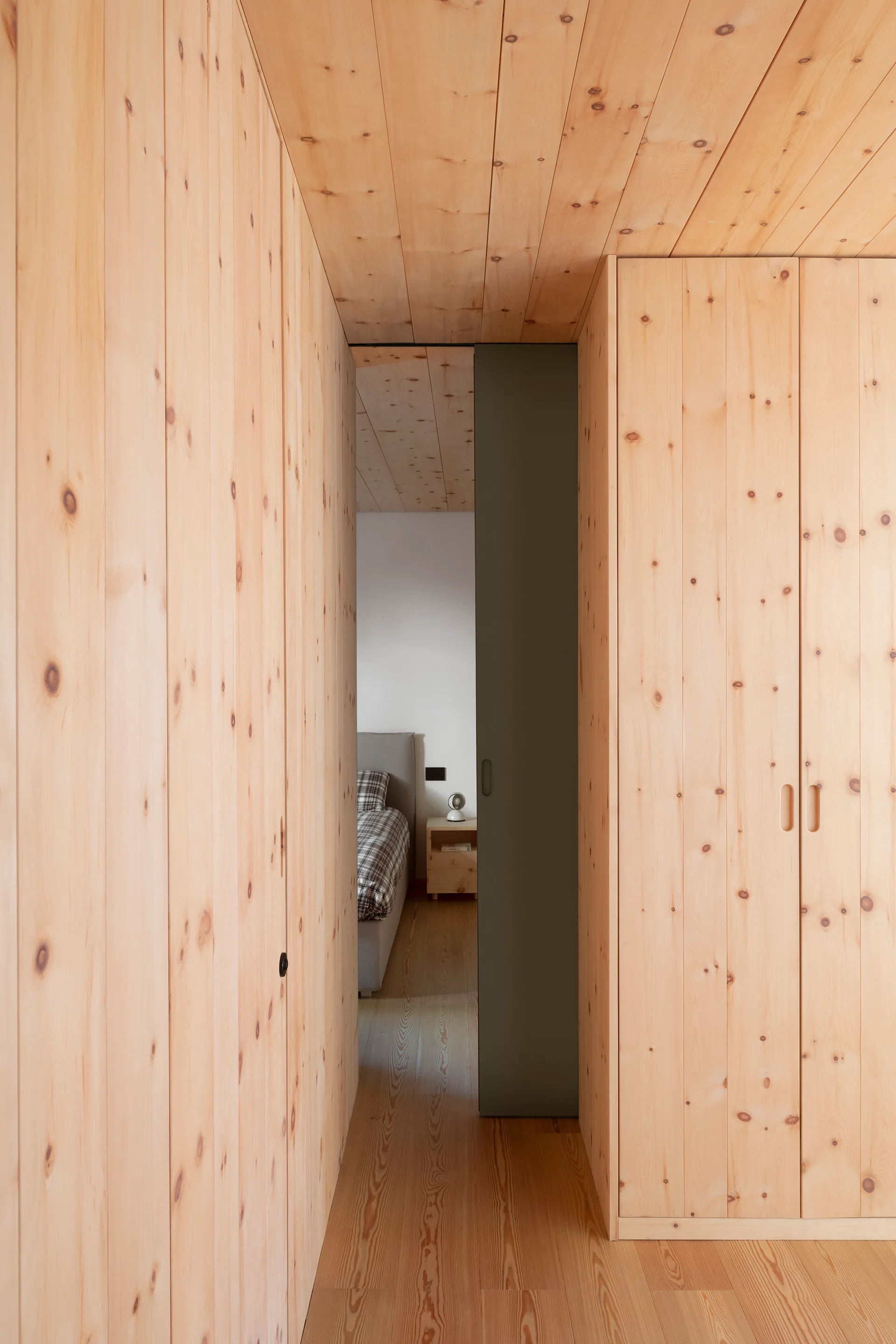 A hallway with knotty pine wood cladding on the walls and ceiling features a dark green sliding door opening into a bedroom.