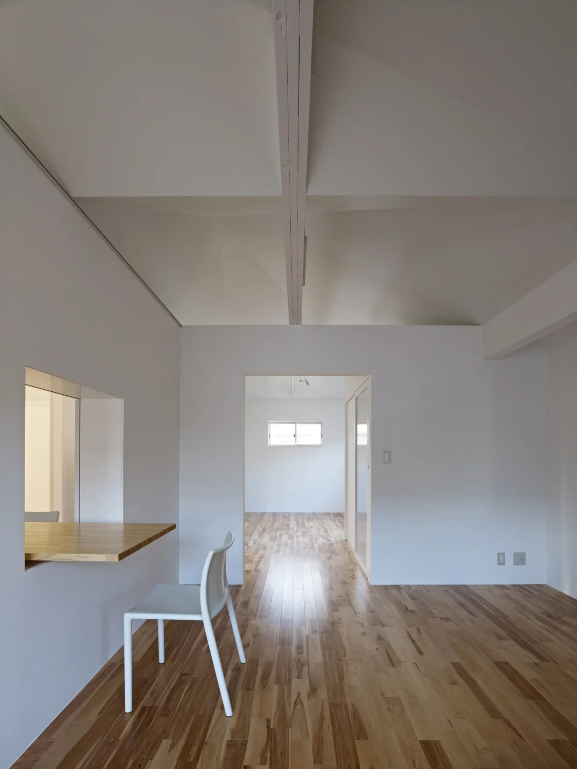 A minimalist white room features natural wood floors, an open doorway, and a built-in wooden counter with a single white plastic chair.