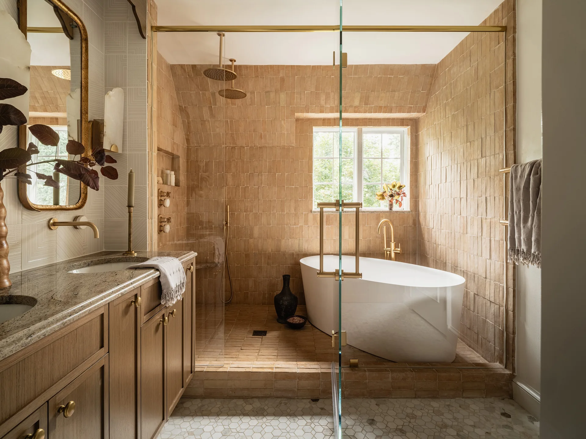 A bathroom wet room features warm tan tiles, a white freestanding bathtub, dual brass shower heads, a glass door, and a wooden vanity.
