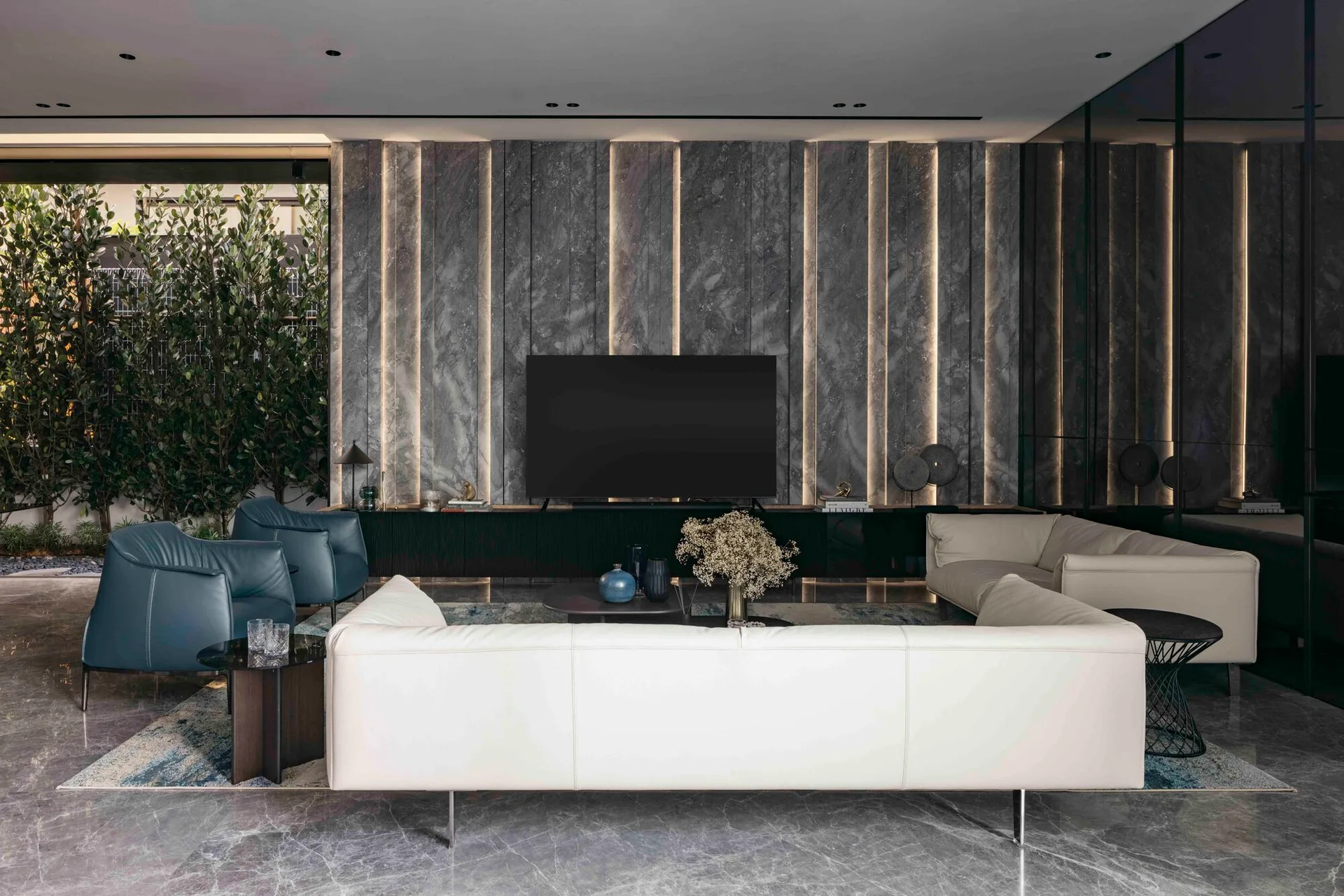 A contemporary living room featuring a white sofa, two blue armchairs, and a grey stone-clad feature wall with vertical integrated accent lighting.