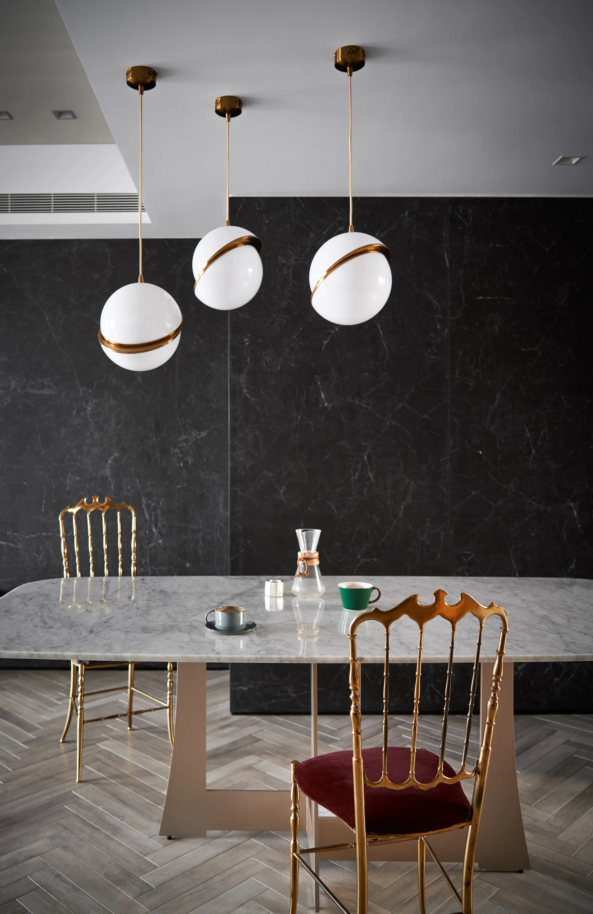 A dining room features a white marble table, ornate gold chairs with dark red seats, and three hanging globe pendant lights against a black marble wall.