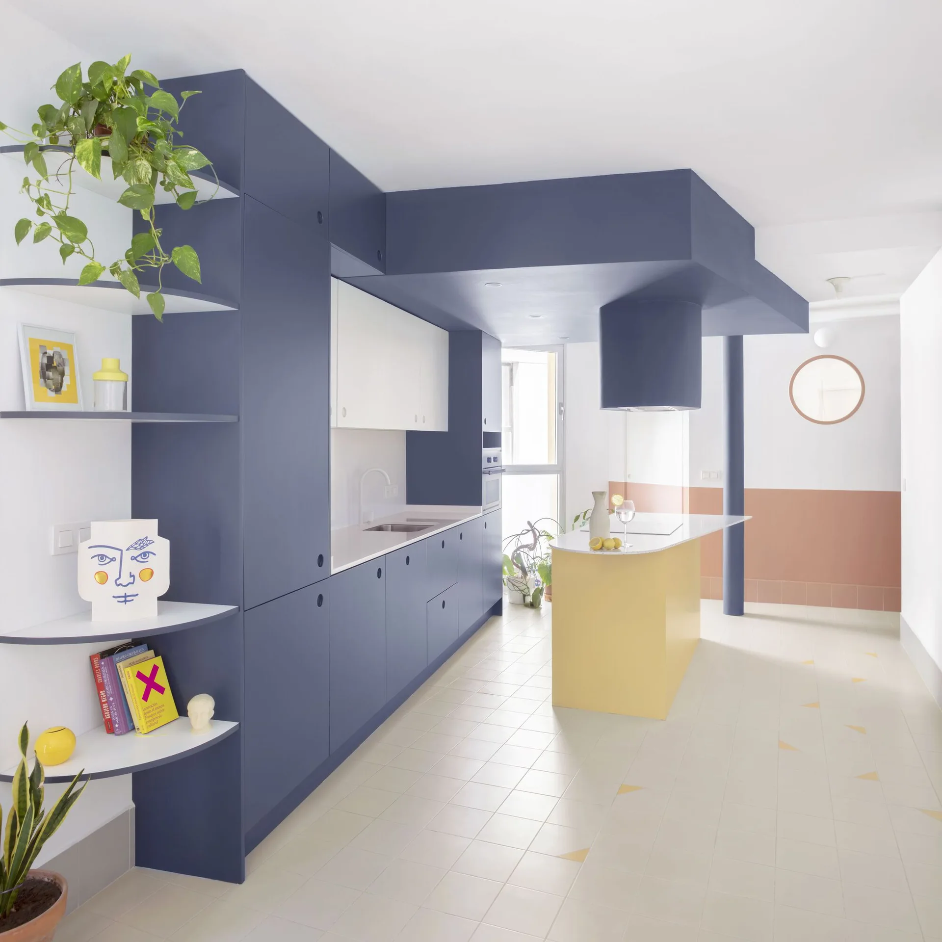 A modern kitchen featuring matte blue cabinetry with curved open shelves, a yellow island with white countertop, and soft pink and white walls.