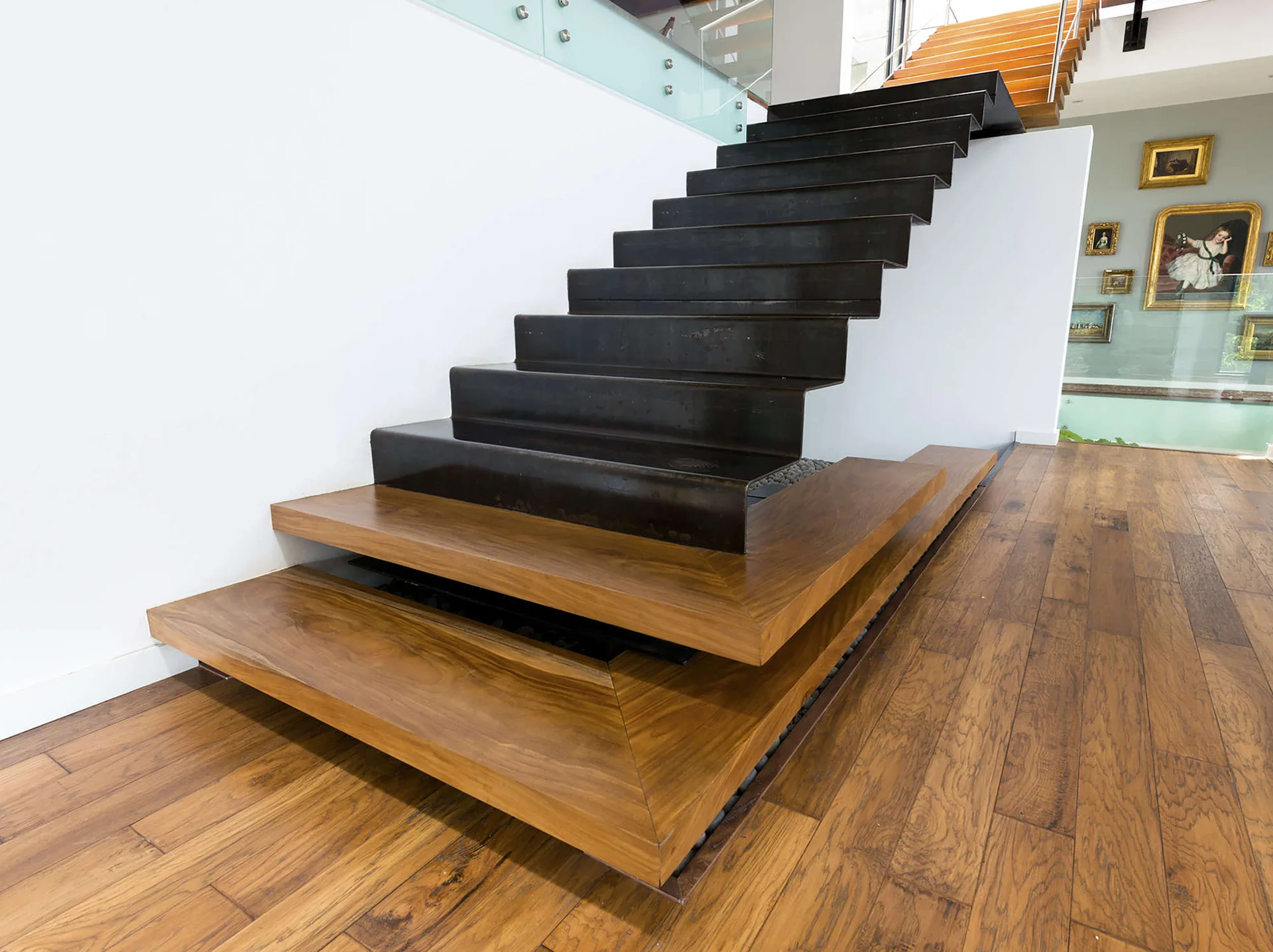A modern staircase features dark folded metal steps rising from a tiered wood-toned platform base onto a hardwood floor against white walls.