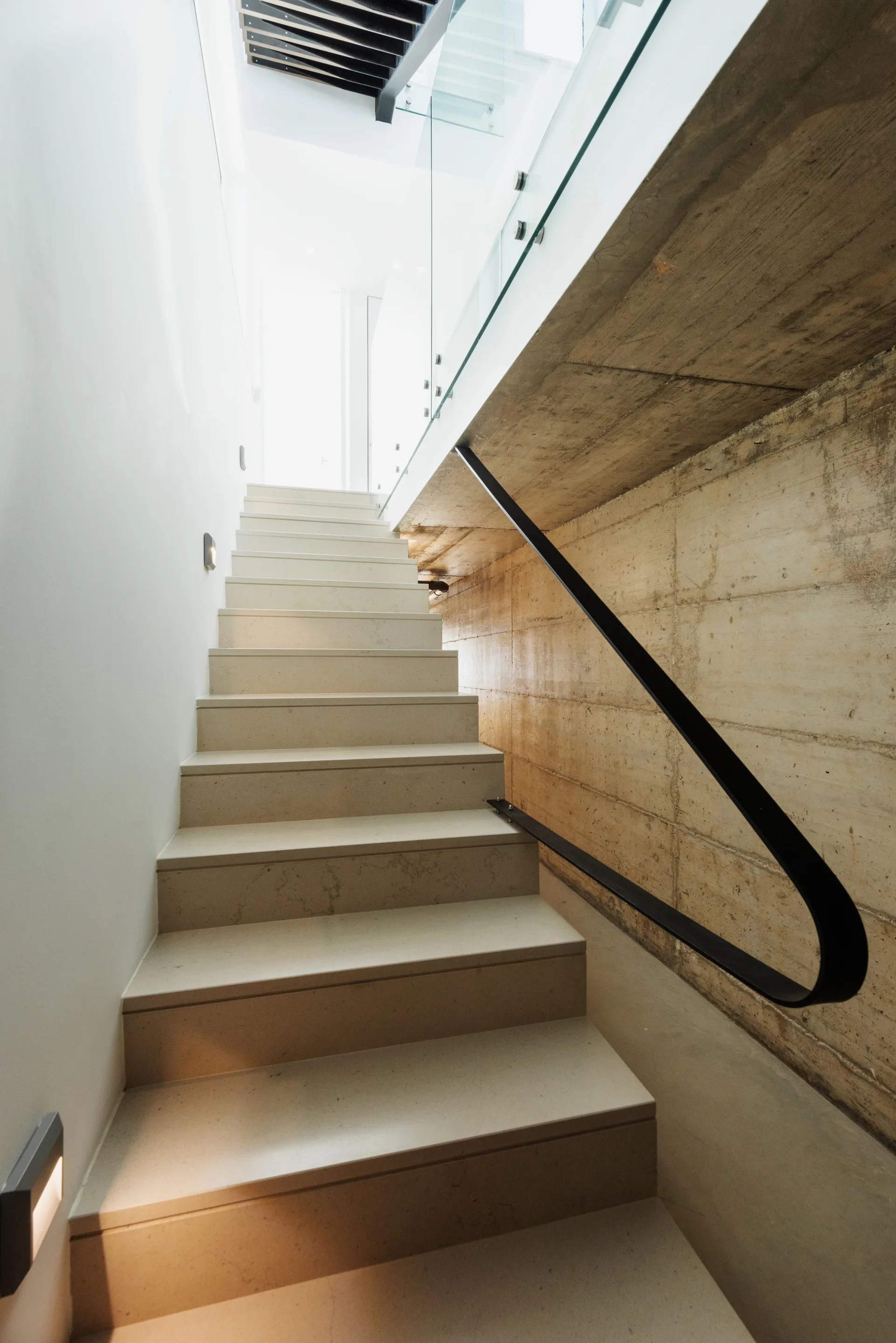 A modern interior staircase with beige stone treads stands beside a textured concrete wall, featuring a black metal handrail and a glass railing.