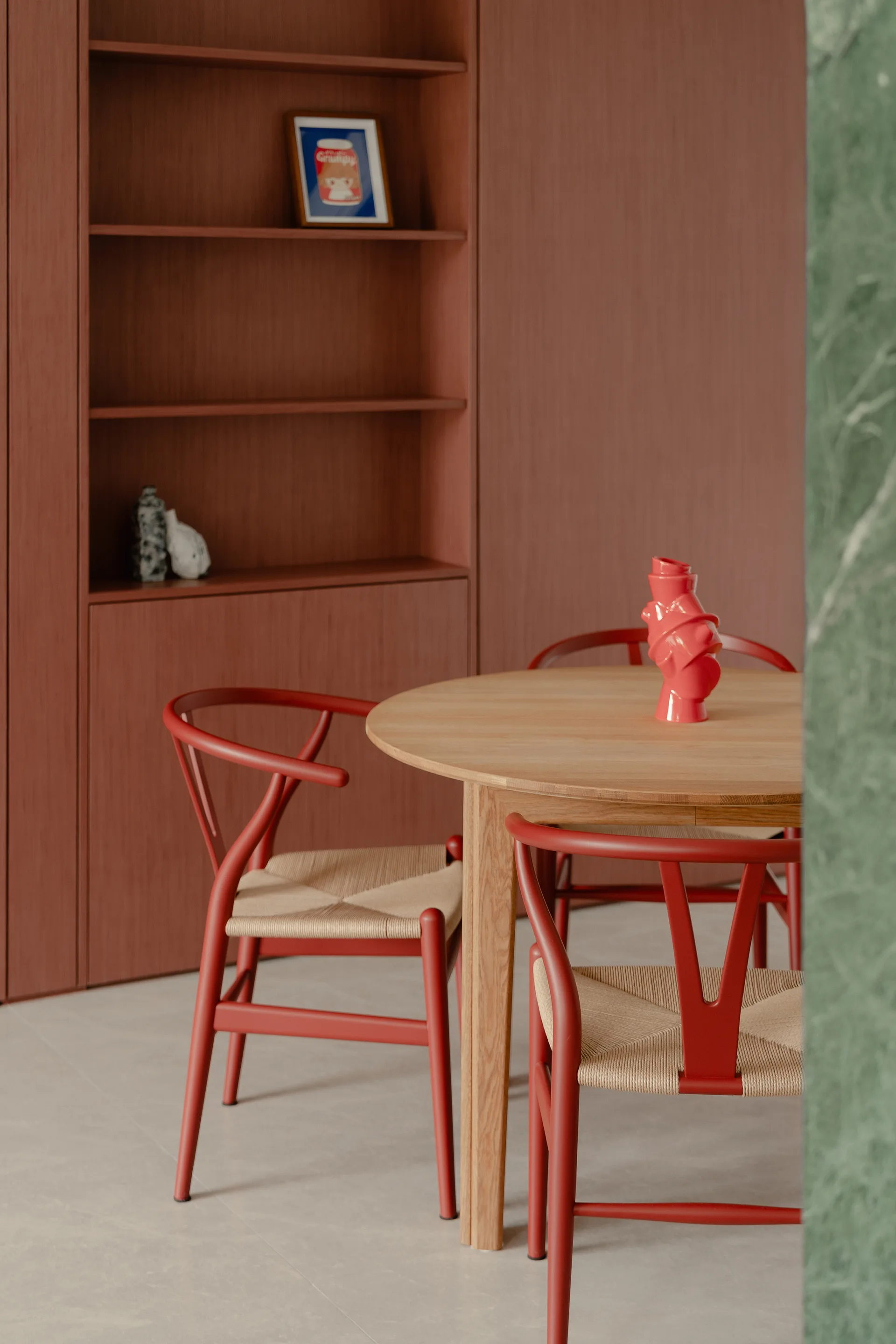 A round wooden dining table is paired with red wishbone chairs with woven seats against a terracotta-colored built-in shelving wall.