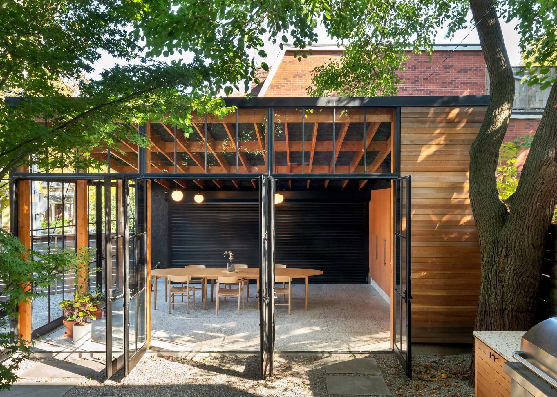 A modern outdoor dining pavilion with a wood table and chairs, exposed ceiling beams, and black-framed glass folding doors opened to a garden.