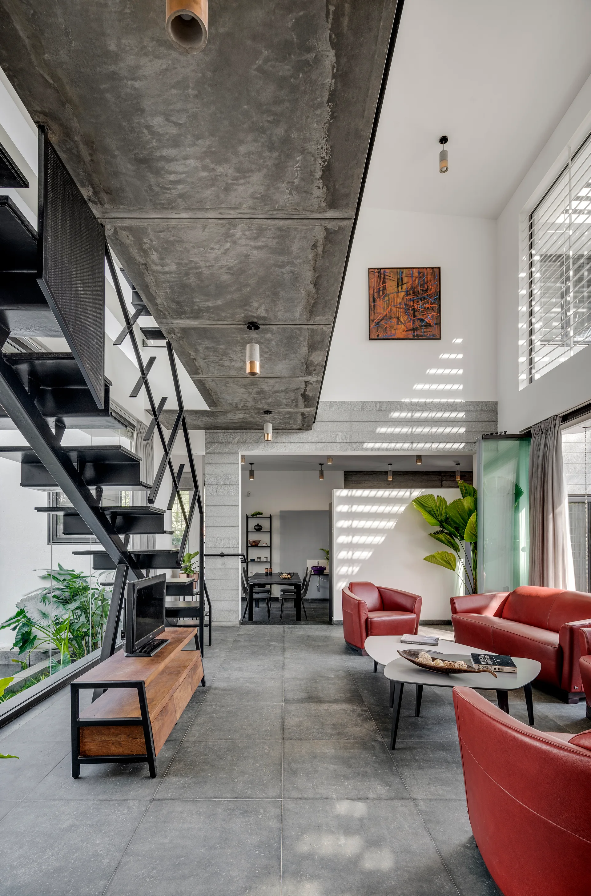 An industrial living room features a black metal staircase, a raw concrete ceiling structure, grey tiled floors, and red leather seating.