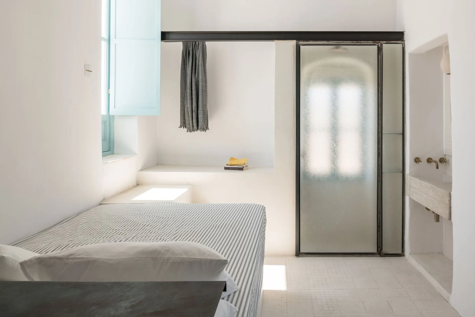 A minimalist white bedroom features a striped bed, a frosted glass partition, pale blue window shutters, and a wall-mounted stone sink.