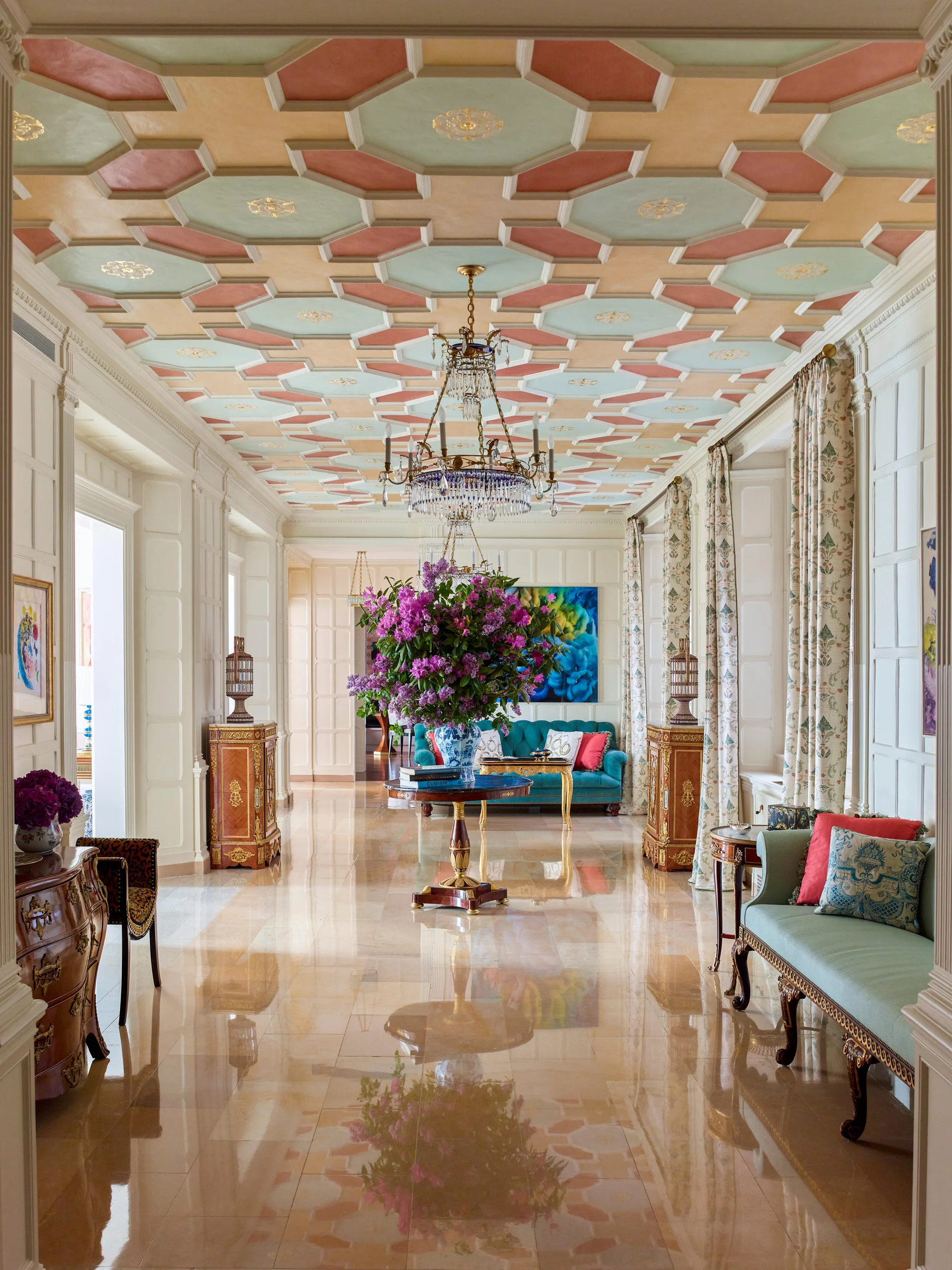 A grand foyer with a pink and green geometric coffered ceiling, polished stone flooring, and a large central purple floral arrangement.