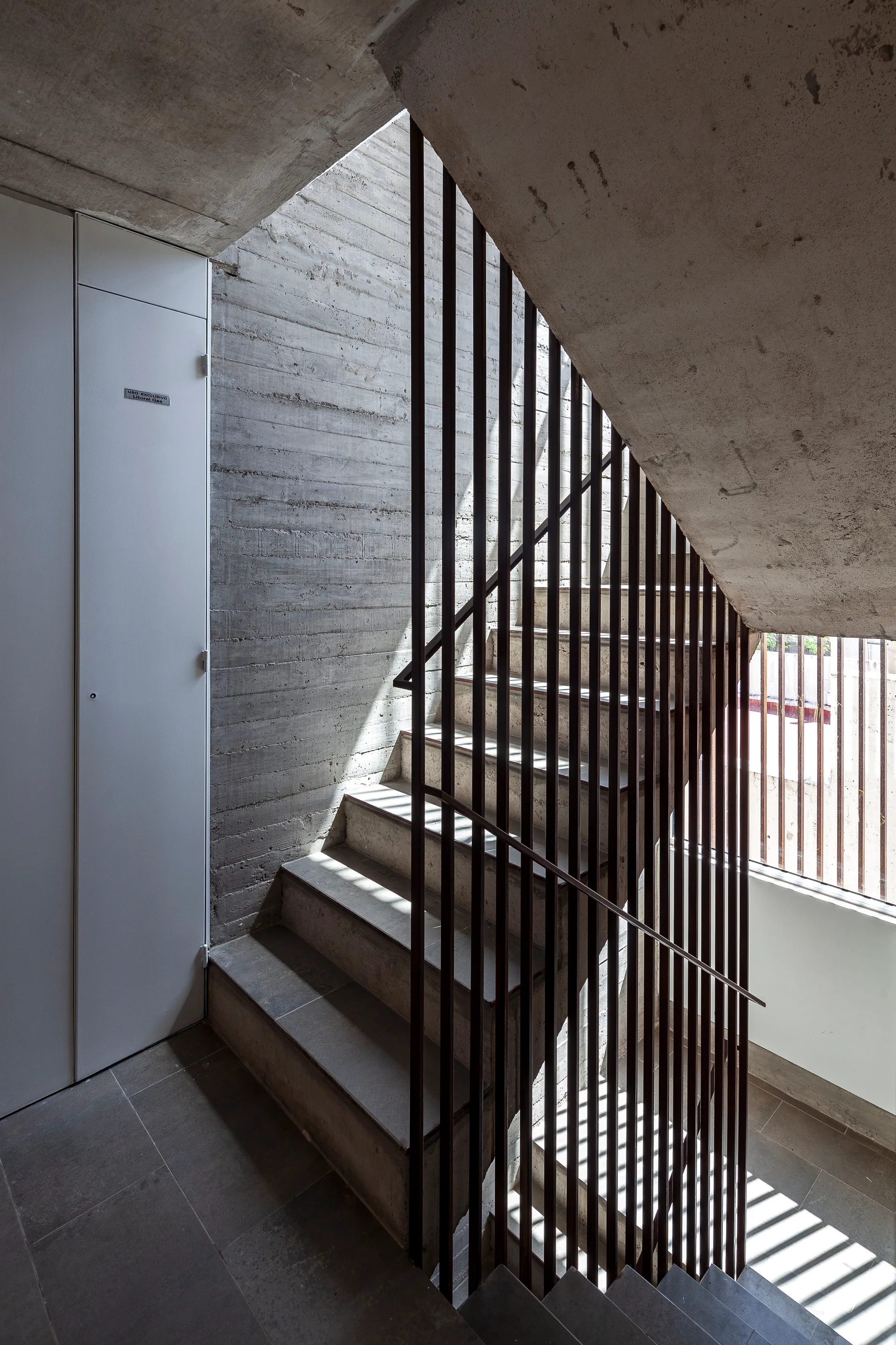 A modern concrete staircase features textured board-marked concrete walls and a dark vertical rod metal railing with sunlight casting striped shadows.