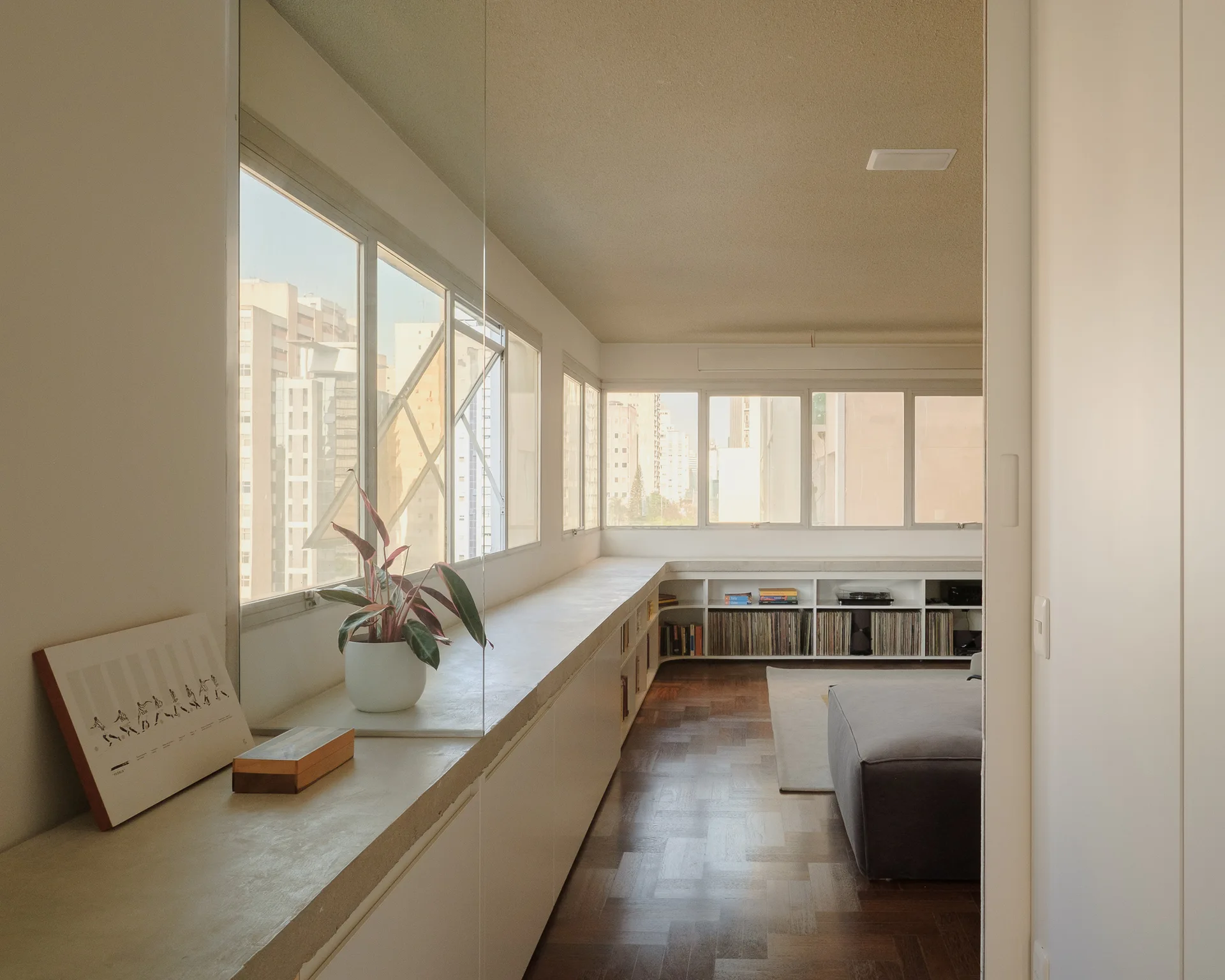 A modernist apartment living room features a long white low-built-in cabinet beneath horizontal windows, a grey sofa, and parquet wood flooring.