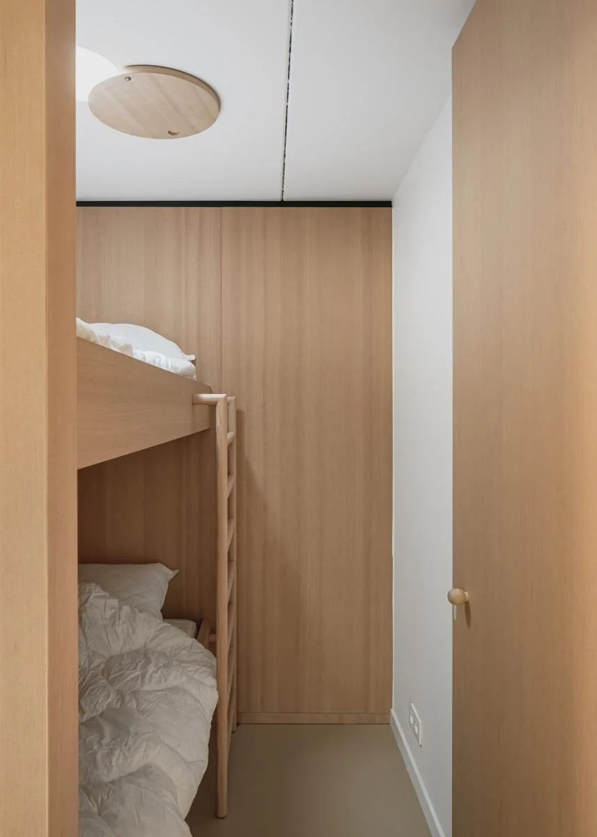 A small minimalist bedroom featuring a built-in wooden bunk bed with white bedding set against a light wood paneled wall.