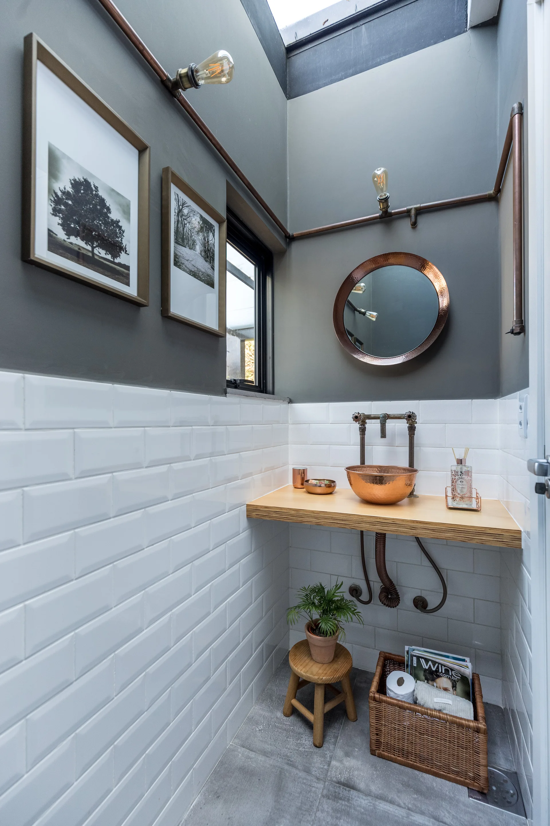 A small powder room with a floating wood vanity, copper vessel sink, round metallic mirror, exposed plumbing, and white subway wall tiles.