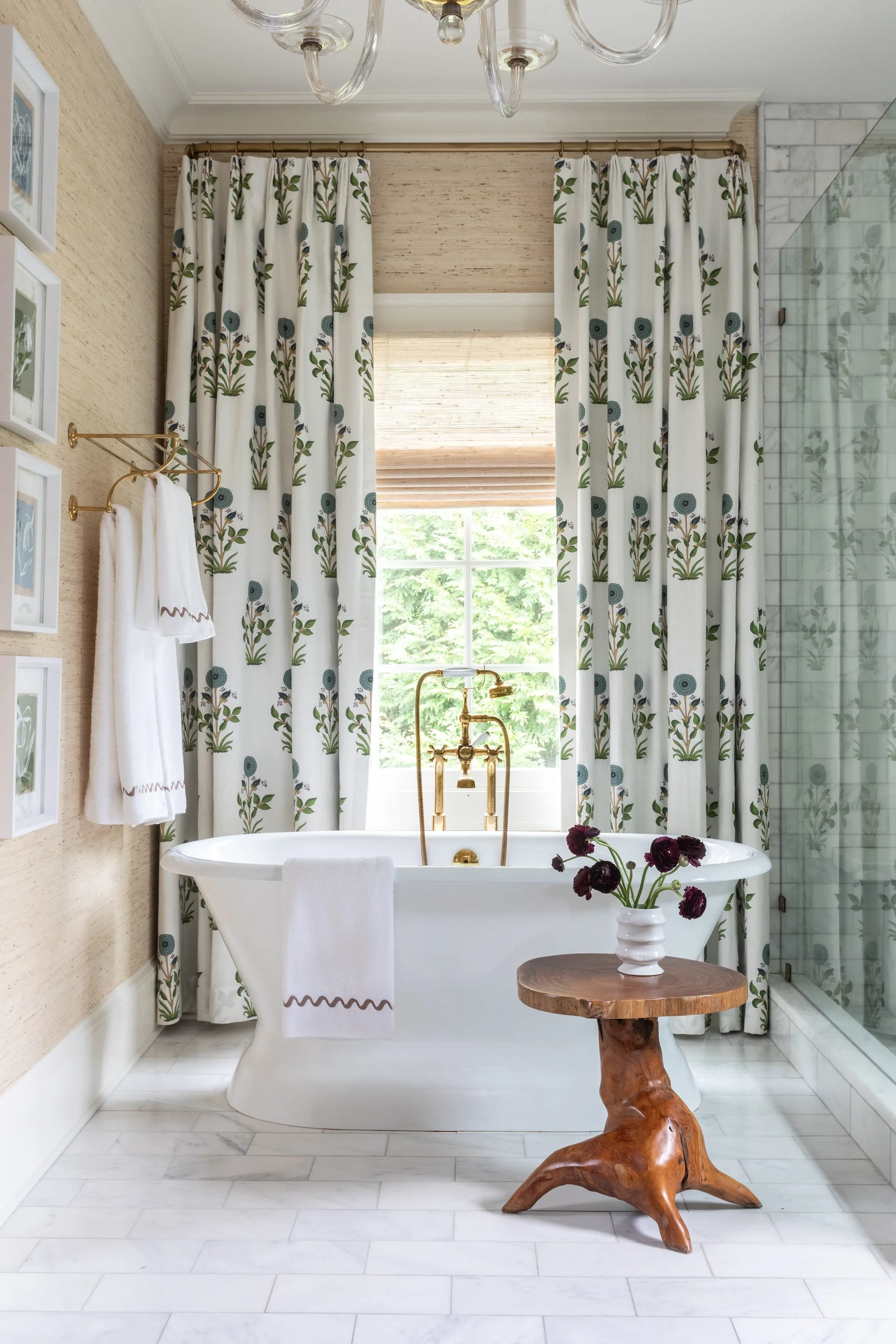 A bright bathroom with a freestanding white bathtub, floral print curtains, a rustic wood side table, and textured grasscloth wallpaper.