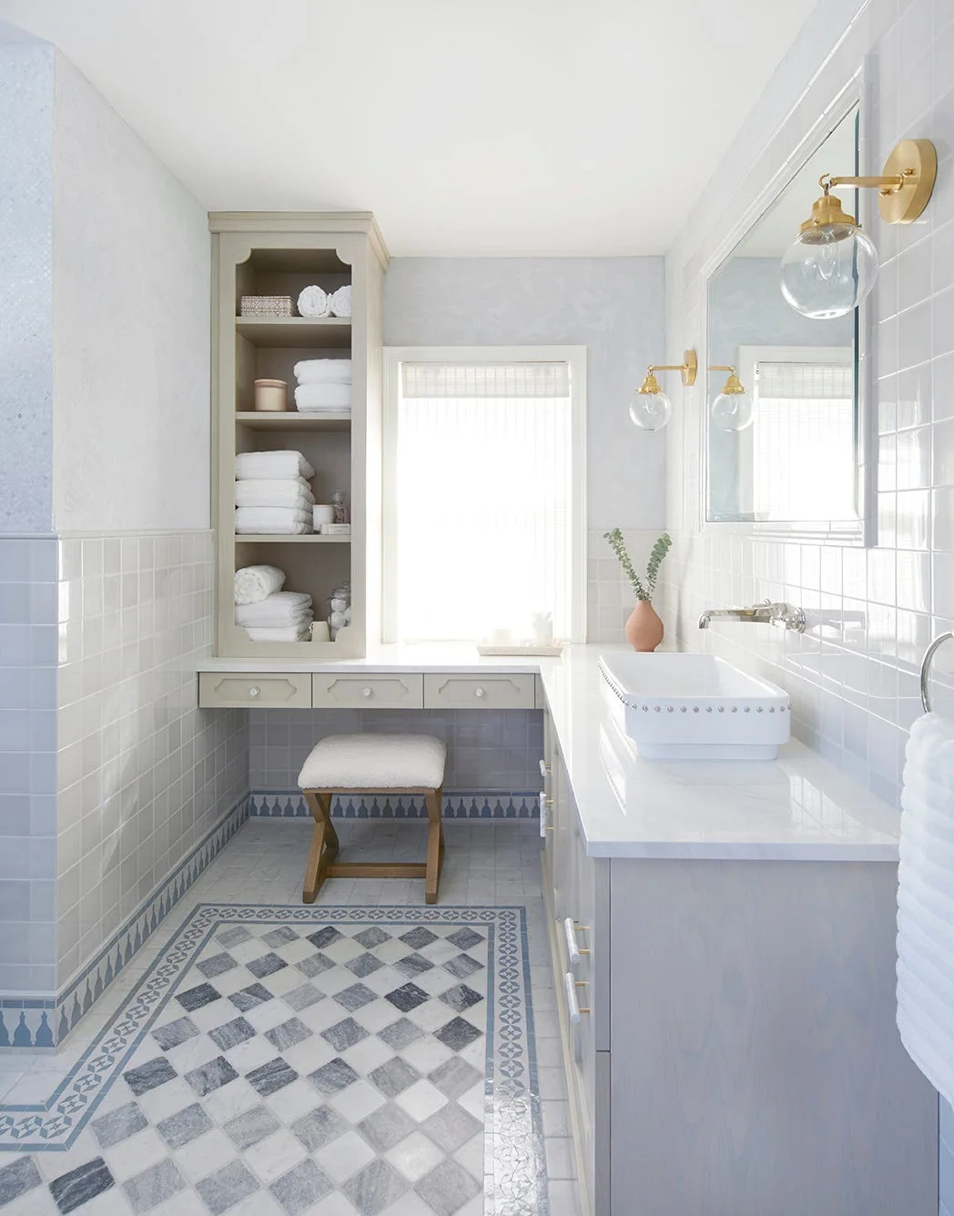 A light blue bathroom features a grey wood vanity with brass sconces, a tall built-in linen shelf, and a blue and white checkered tiled floor.