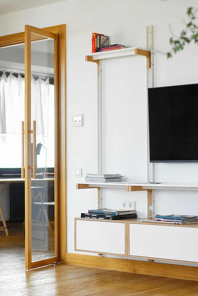 A white wall-mounted modular shelving unit with light wood brackets next to a wood-framed glass door.