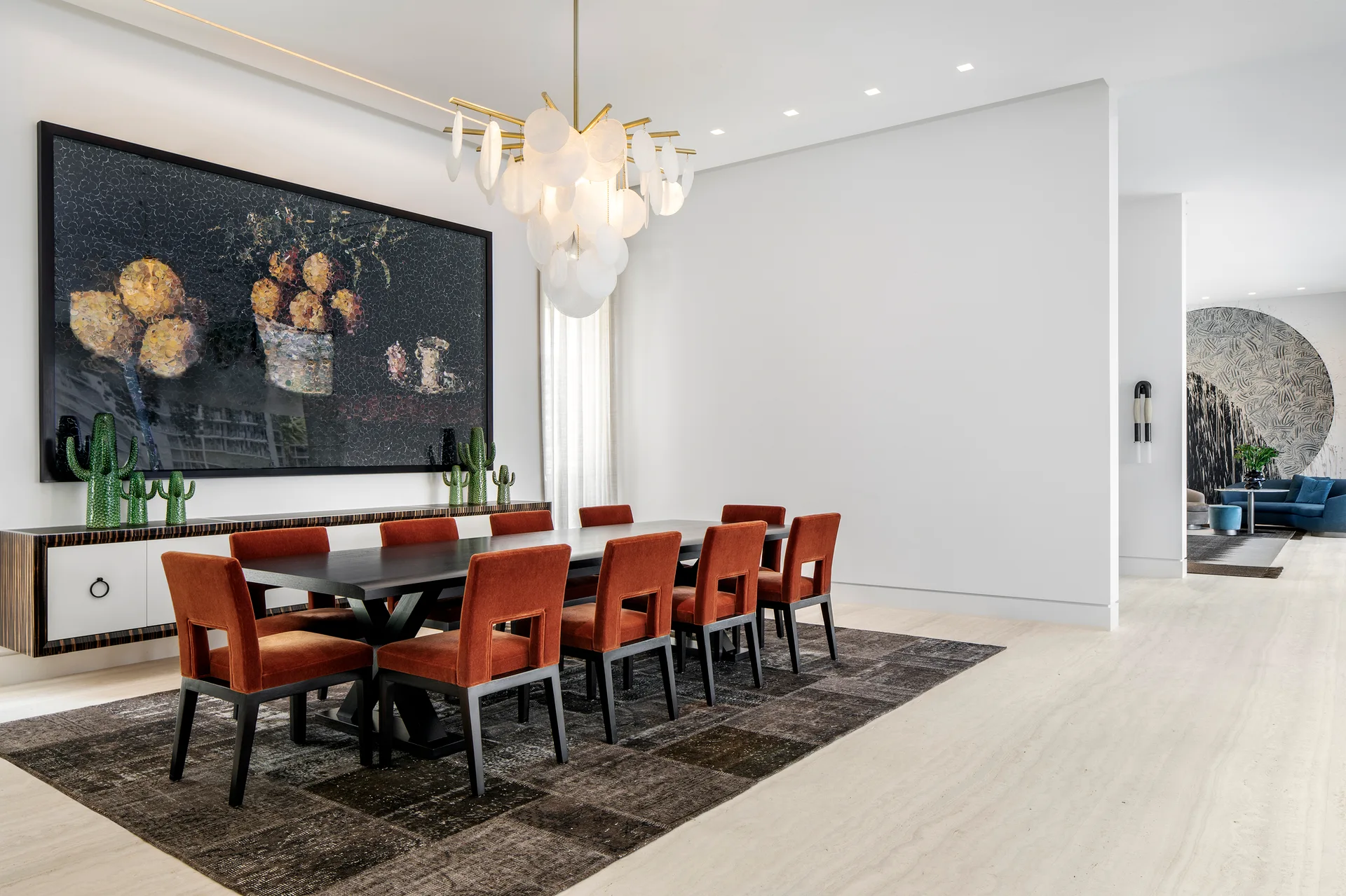 A modern dining room features a dark wood table surrounded by rust-colored chairs beneath a tiered disc chandelier and a large framed painting.
