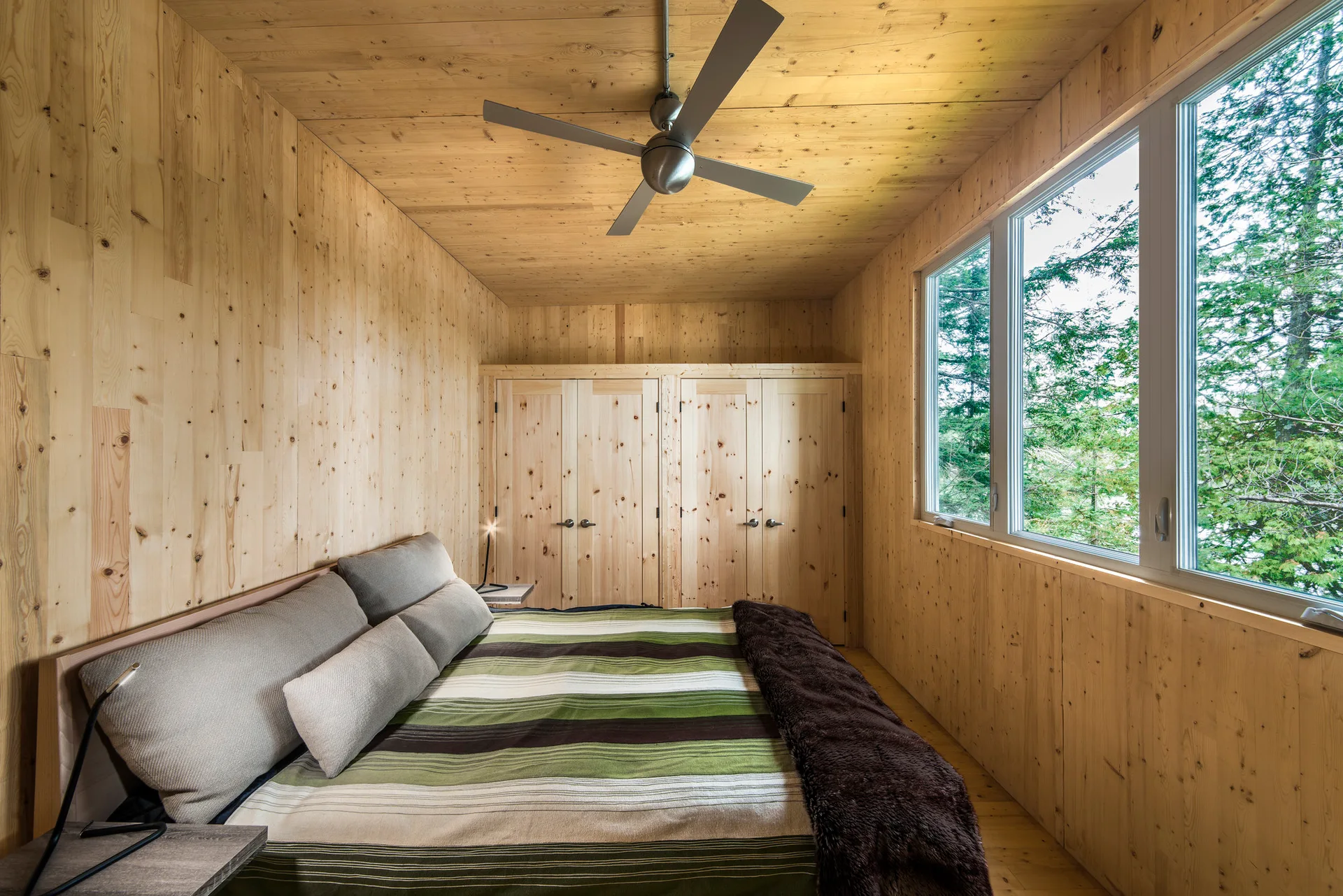 A rustic bedroom with natural wood paneled walls and ceiling features a bed with green striped bedding near a large window.