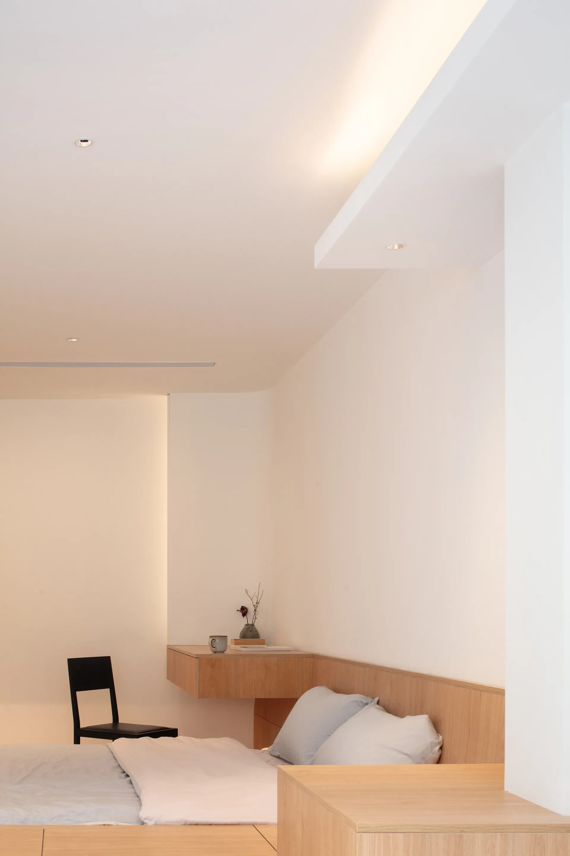 A minimalist bedroom features a floating light wood desk extending from matching wall paneling, lit by soft recessed ceiling and cove lights.