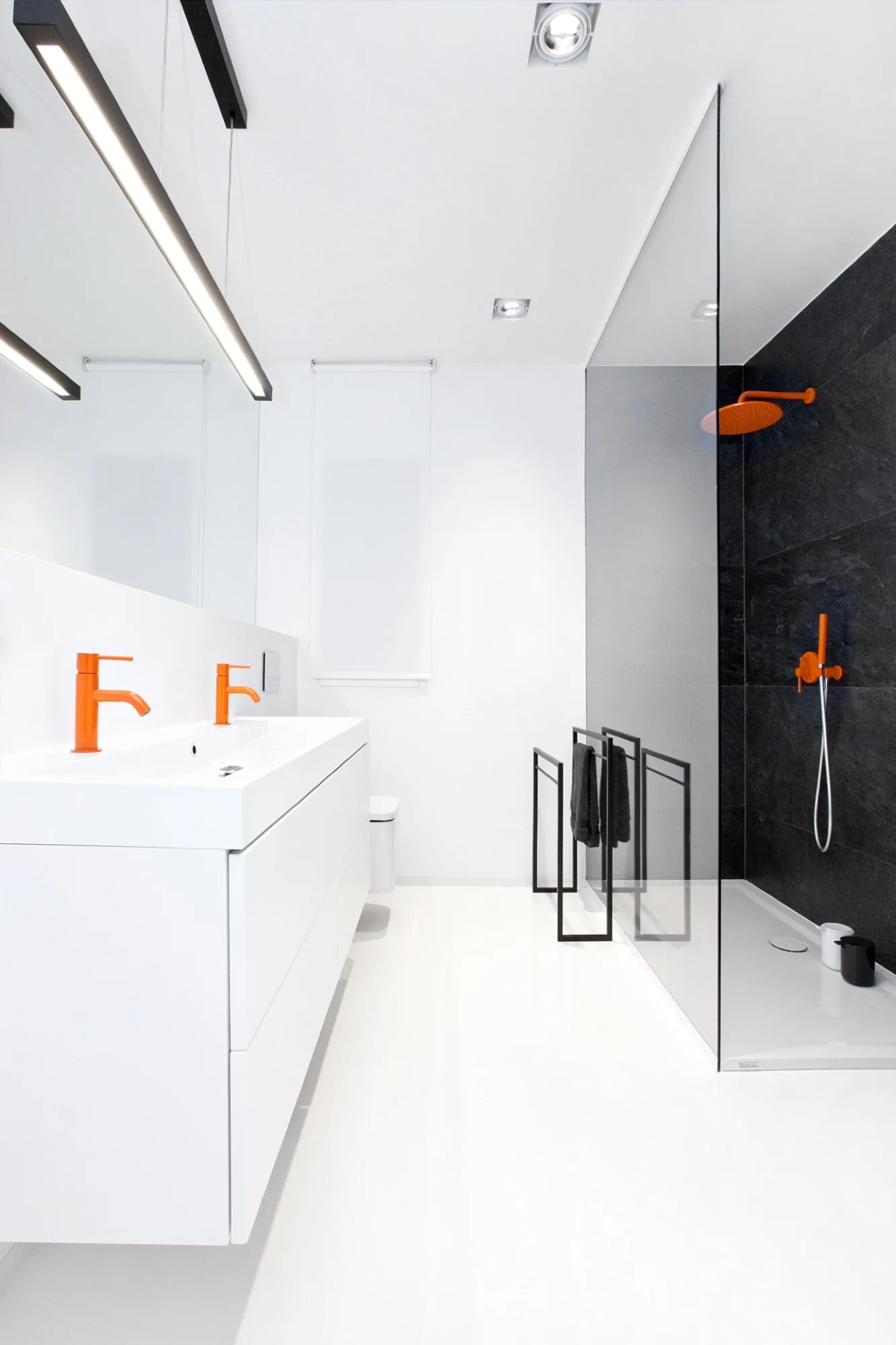 A minimalist white bathroom features a double vanity with bright orange faucets next to a walk-in shower with black tile walls.