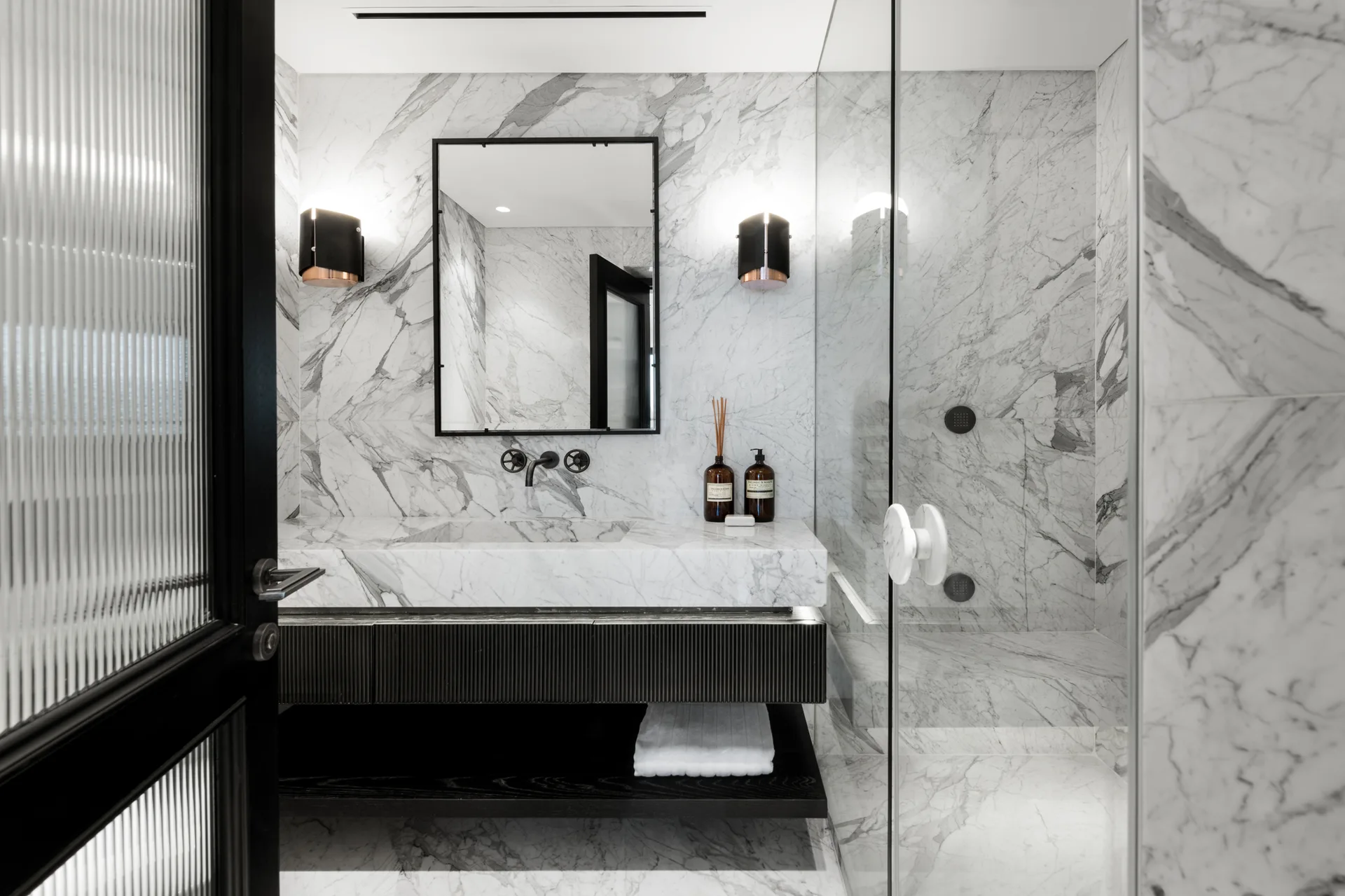 A modern bathroom featuring veined white marble walls and floors, a floating vanity with a ribbed black cabinet, and a glass-enclosed shower.