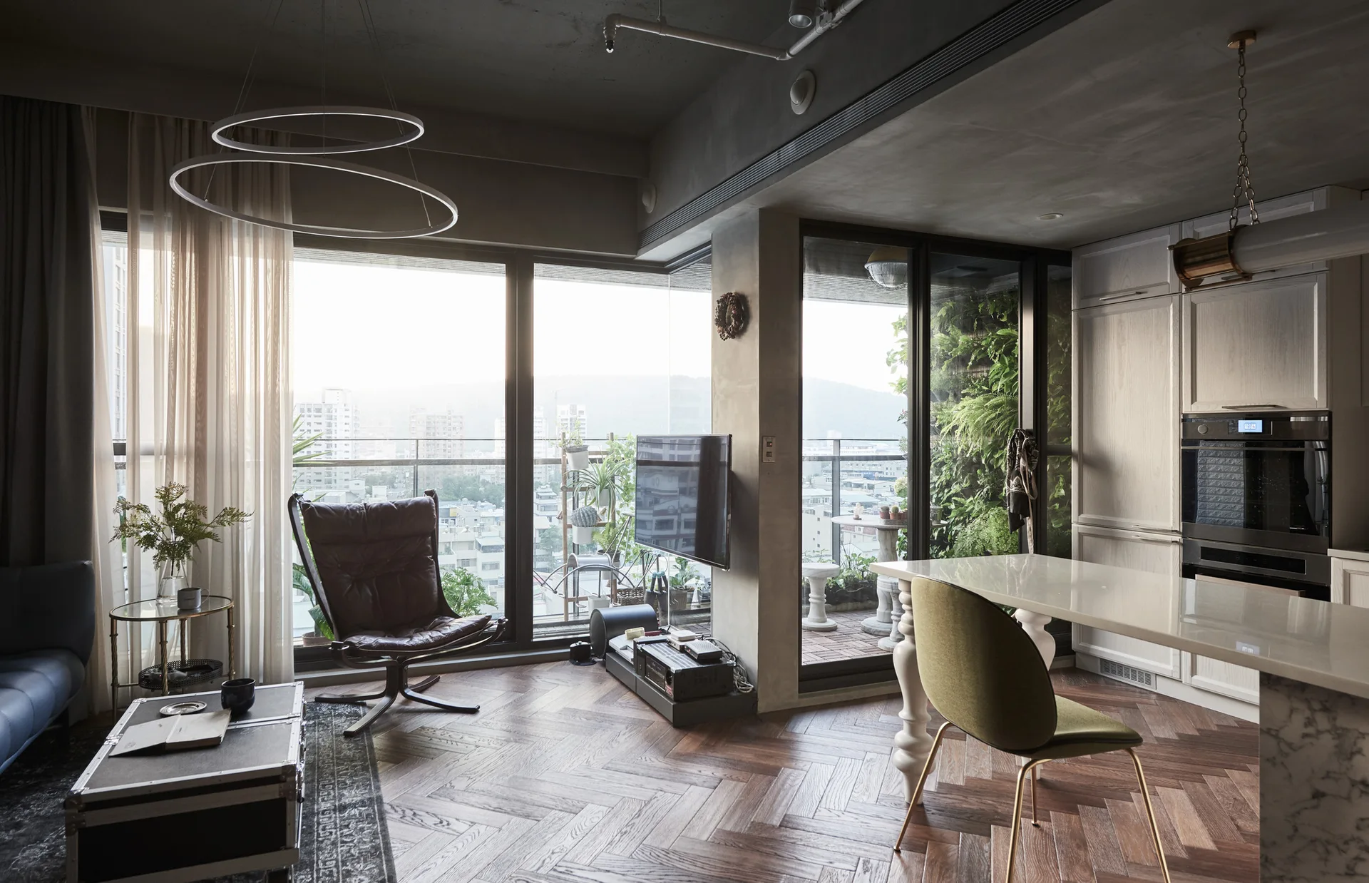 A modern open-plan living room with dark herringbone wood flooring, a grey concrete ceiling, a brown leather armchair, and large windows.