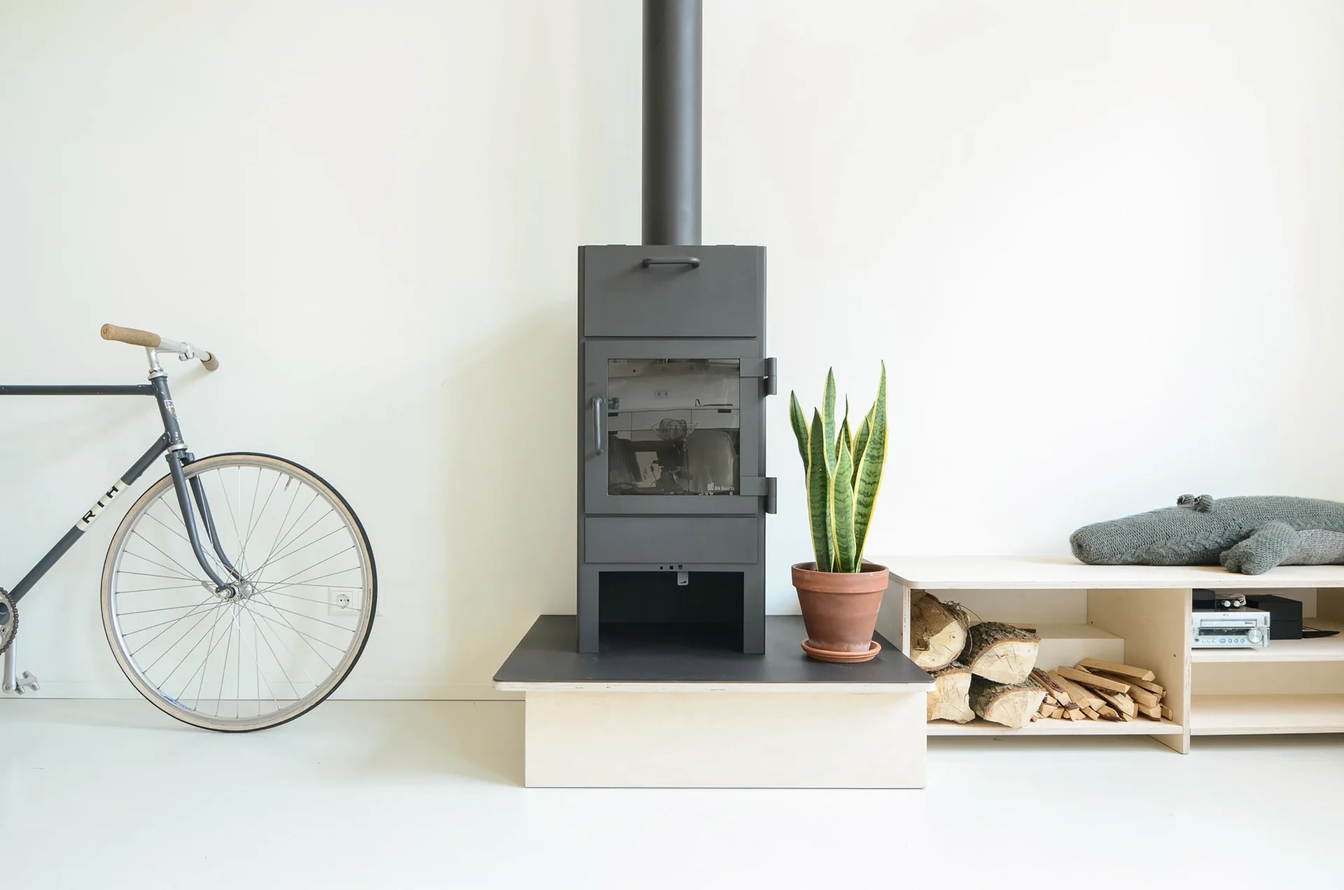 A black wood-burning stove on a raised platform next to a low plywood storage bench with logs, a terracotta pot, and a bicycle on a white floor.