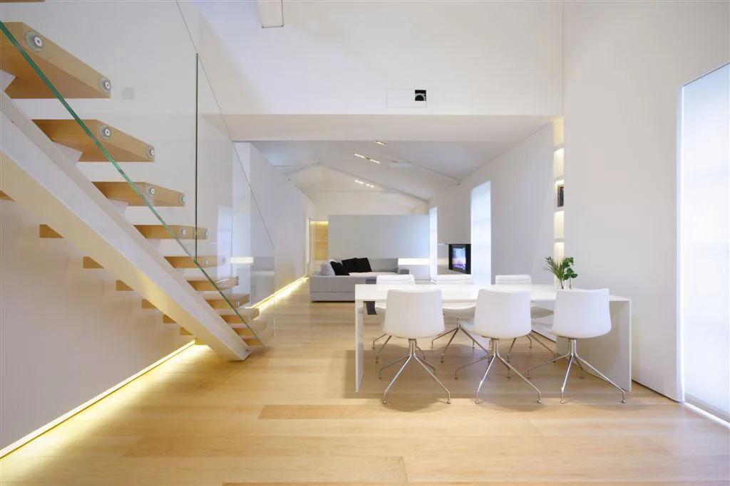 A bright minimalist dining area features a white table and chairs next to a floating wooden staircase with a glass railing and light-colored wood flooring.