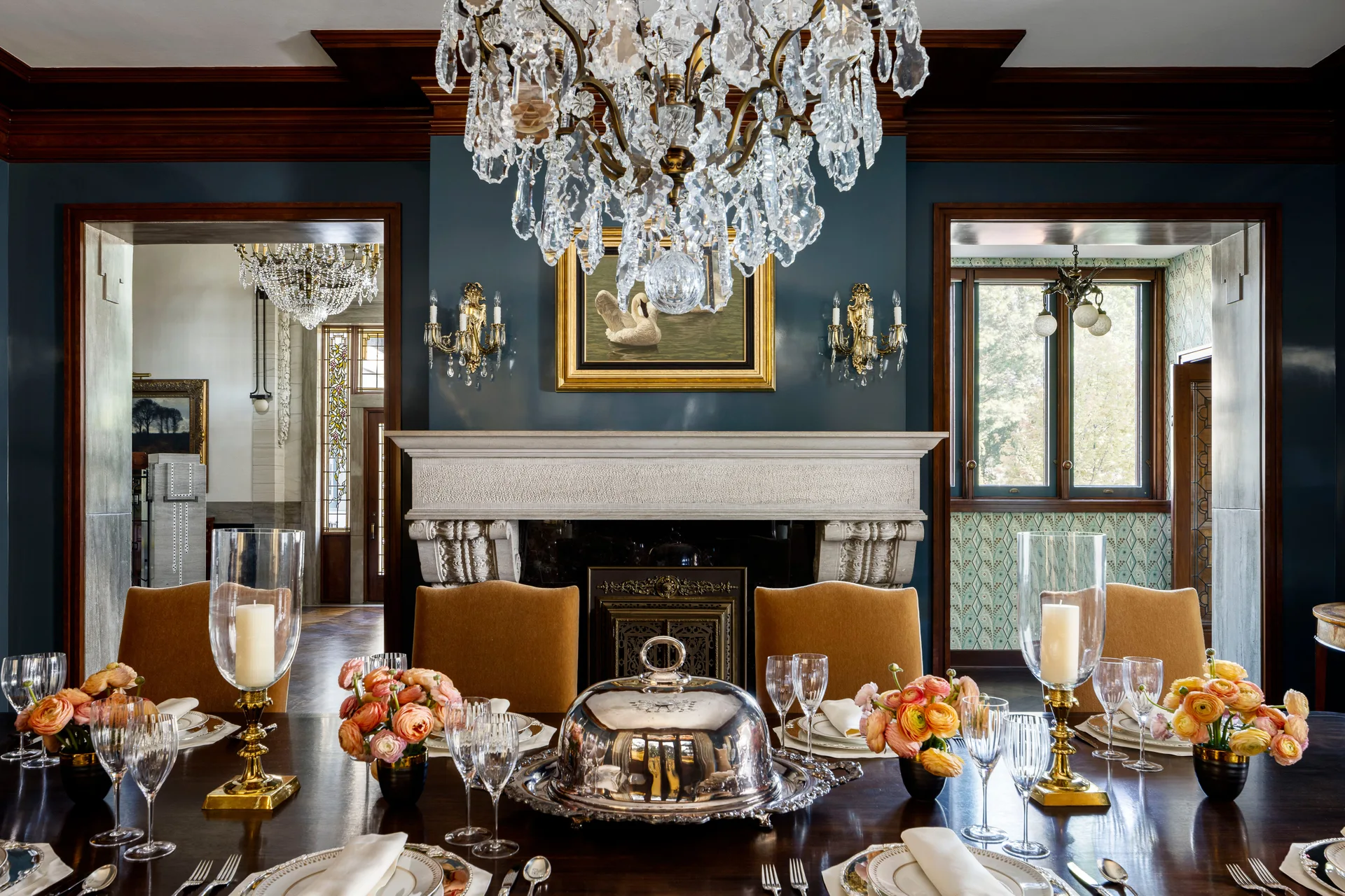 A formal dining table set with silver and florals in front of a stone fireplace, framed by deep teal walls and a crystal chandelier.
