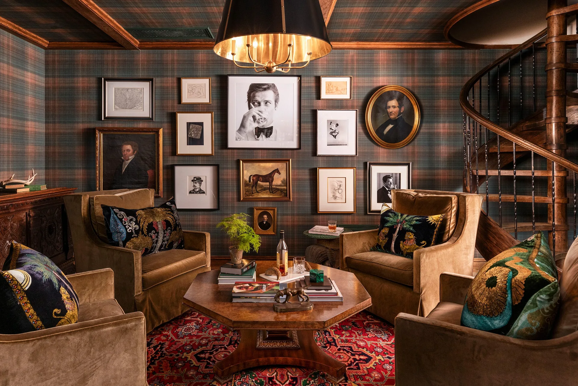 Four brown velvet armchairs surround an octagonal wooden coffee table in a room with tartan plaid wallpaper and a gallery wall.