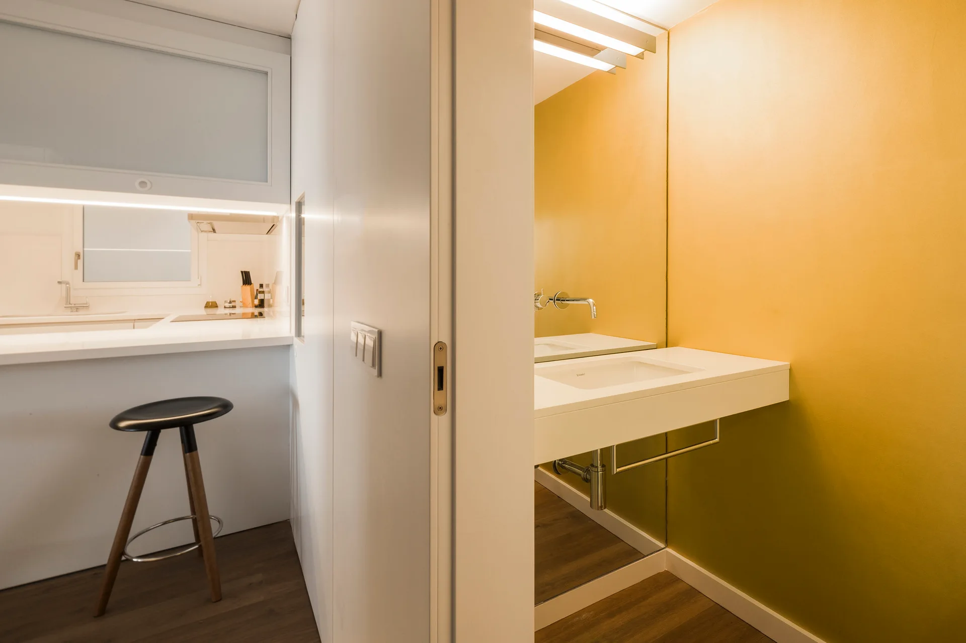 A modern powder room with a vibrant yellow wall and a white floating vanity sink sits adjacent to a clean white kitchen with a bar stool.