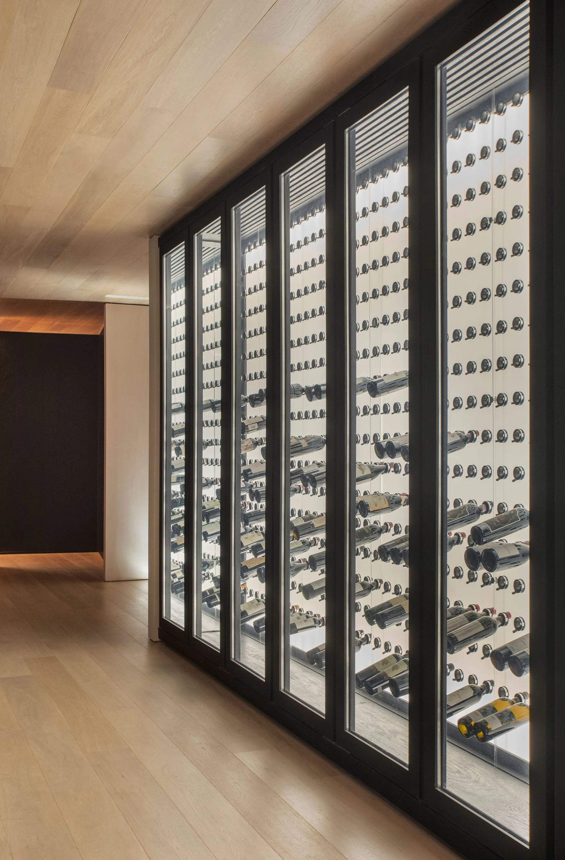 A floor-to-ceiling glass-fronted wine storage unit with a black metal frame set against light wood paneled walls and ceilings.