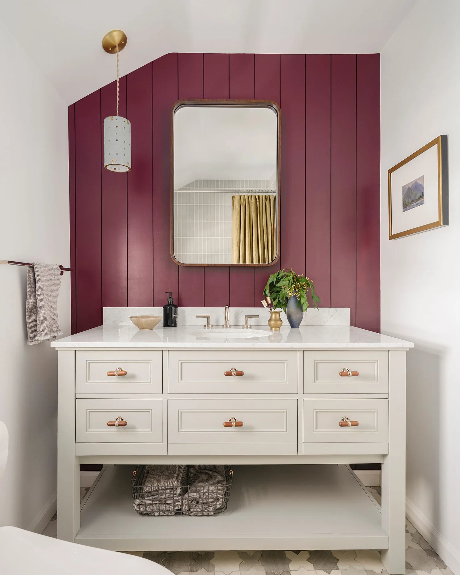 A cream bathroom vanity with a white marble countertop is set against a deep maroon vertical wood paneled wall beneath a rounded rectangular mirror.