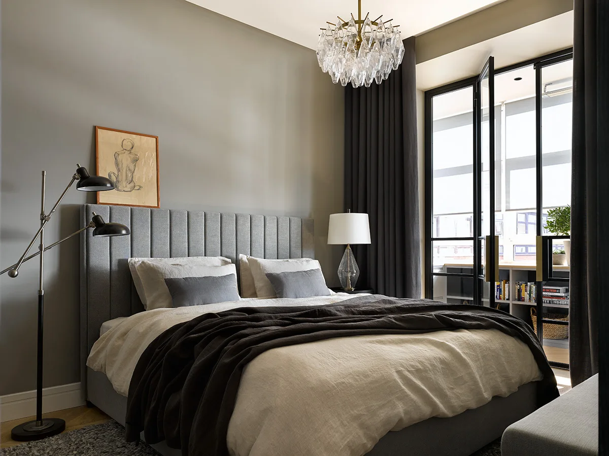 A modern grey bedroom featuring a channel-tufted upholstered bed, a crystal chandelier, and black-framed glass doors leading to an adjacent room.