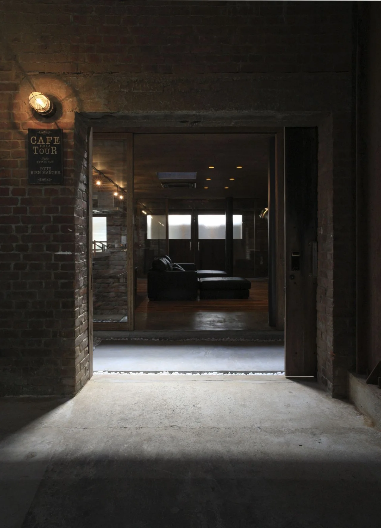 A view through a brick doorway looking into a dark industrial living space with a black leather sofa and warm recessed ceiling lights.