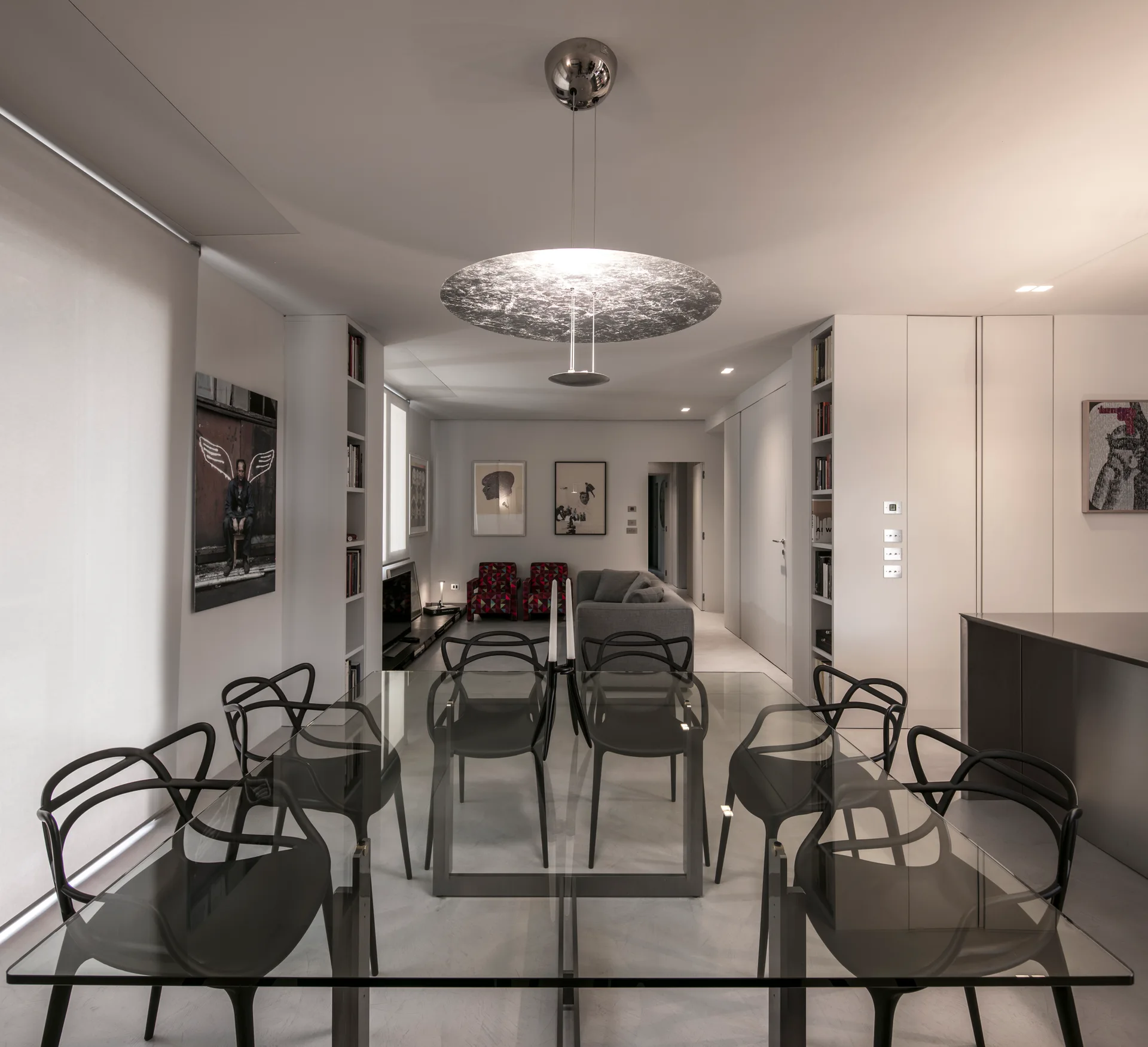 A modern dining room features a glass-topped table surrounded by black curved chairs under a large metallic disc pendant light.