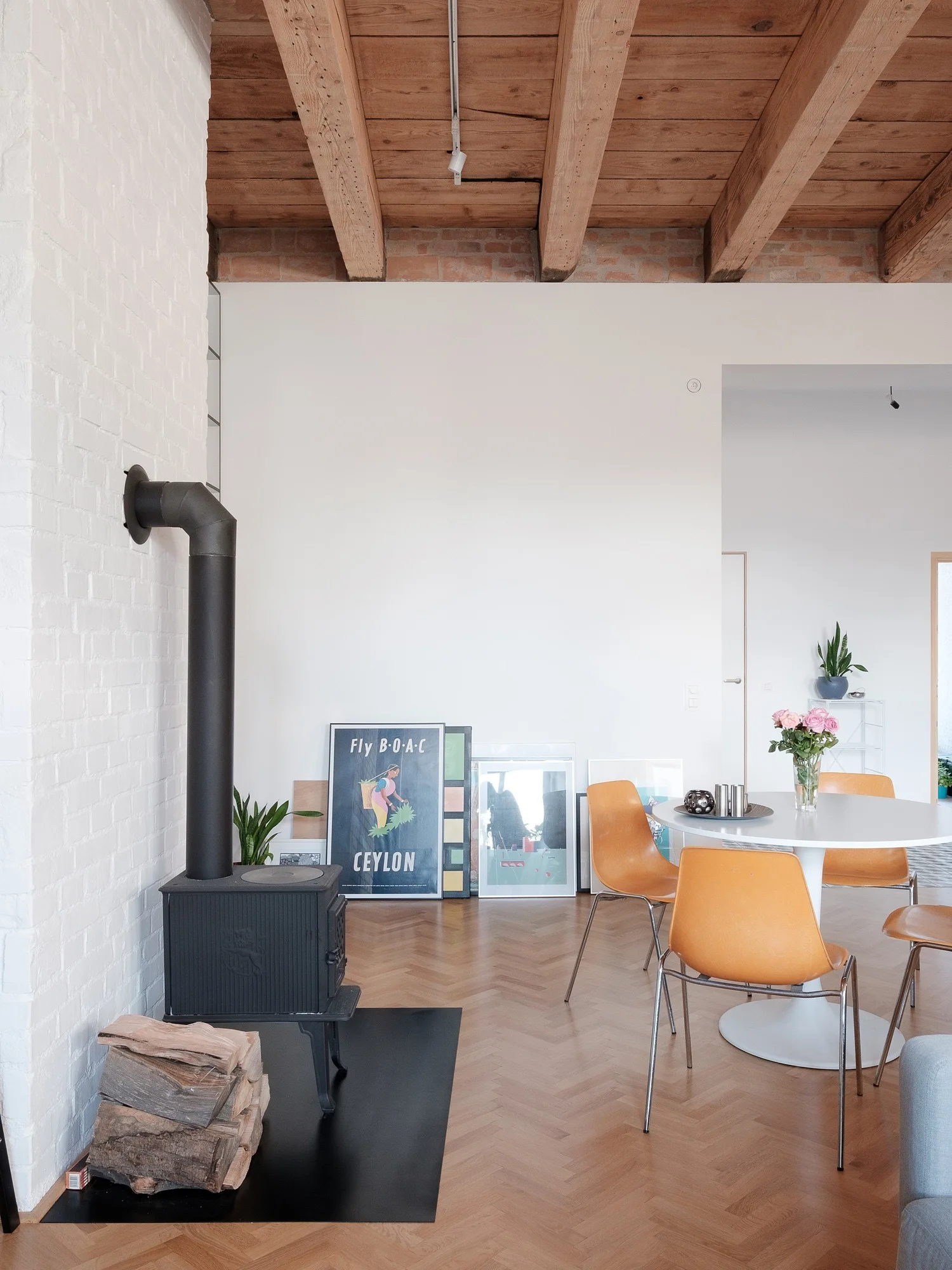 A dining area with a white round pedestal table, orange chairs, a black cast iron stove, herringbone wood floors, and exposed ceiling beams.