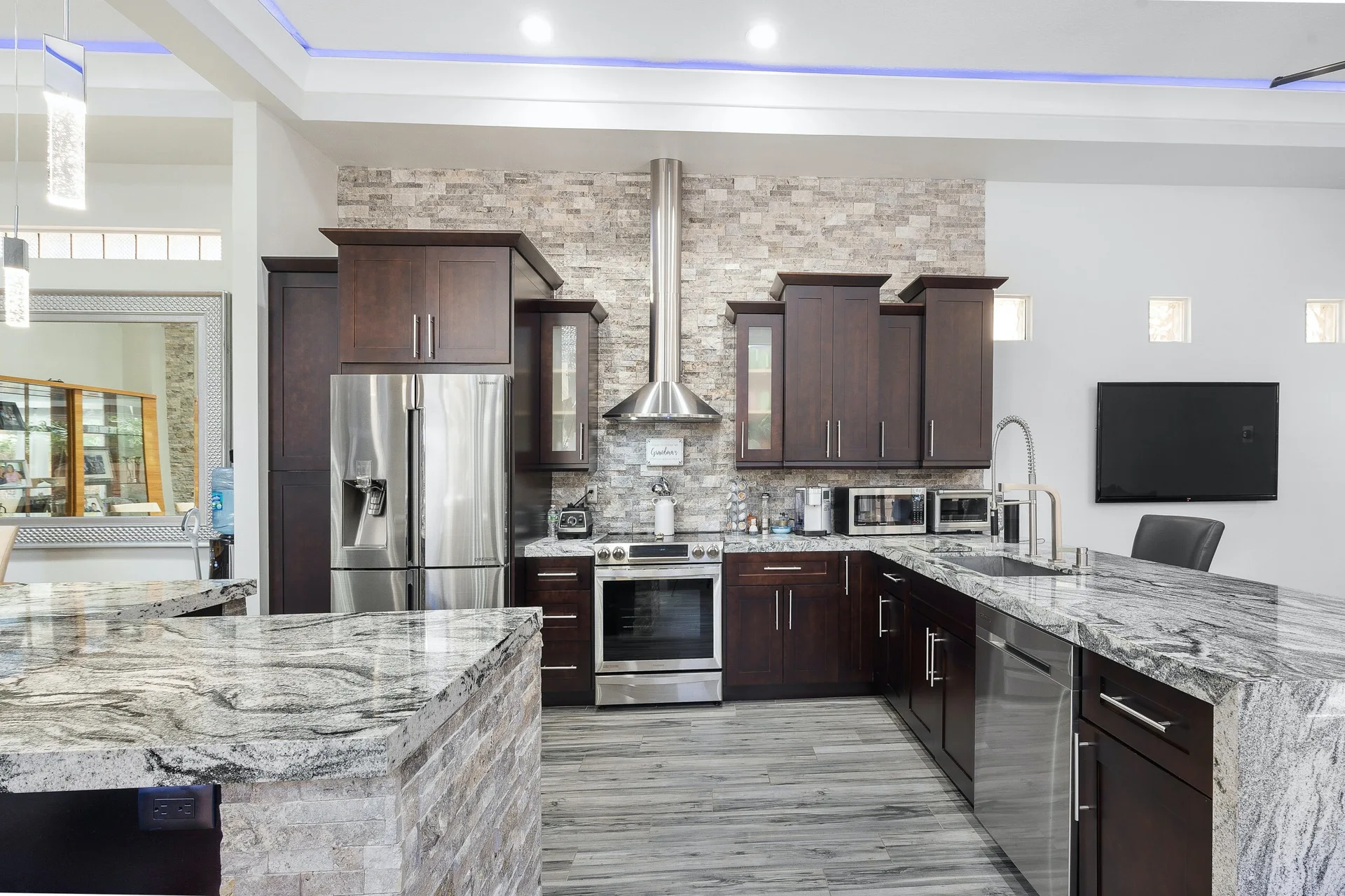 A contemporary kitchen features dark wood cabinets, a stacked stone tile backsplash, heavily veined granite countertops, and stainless steel appliances.
