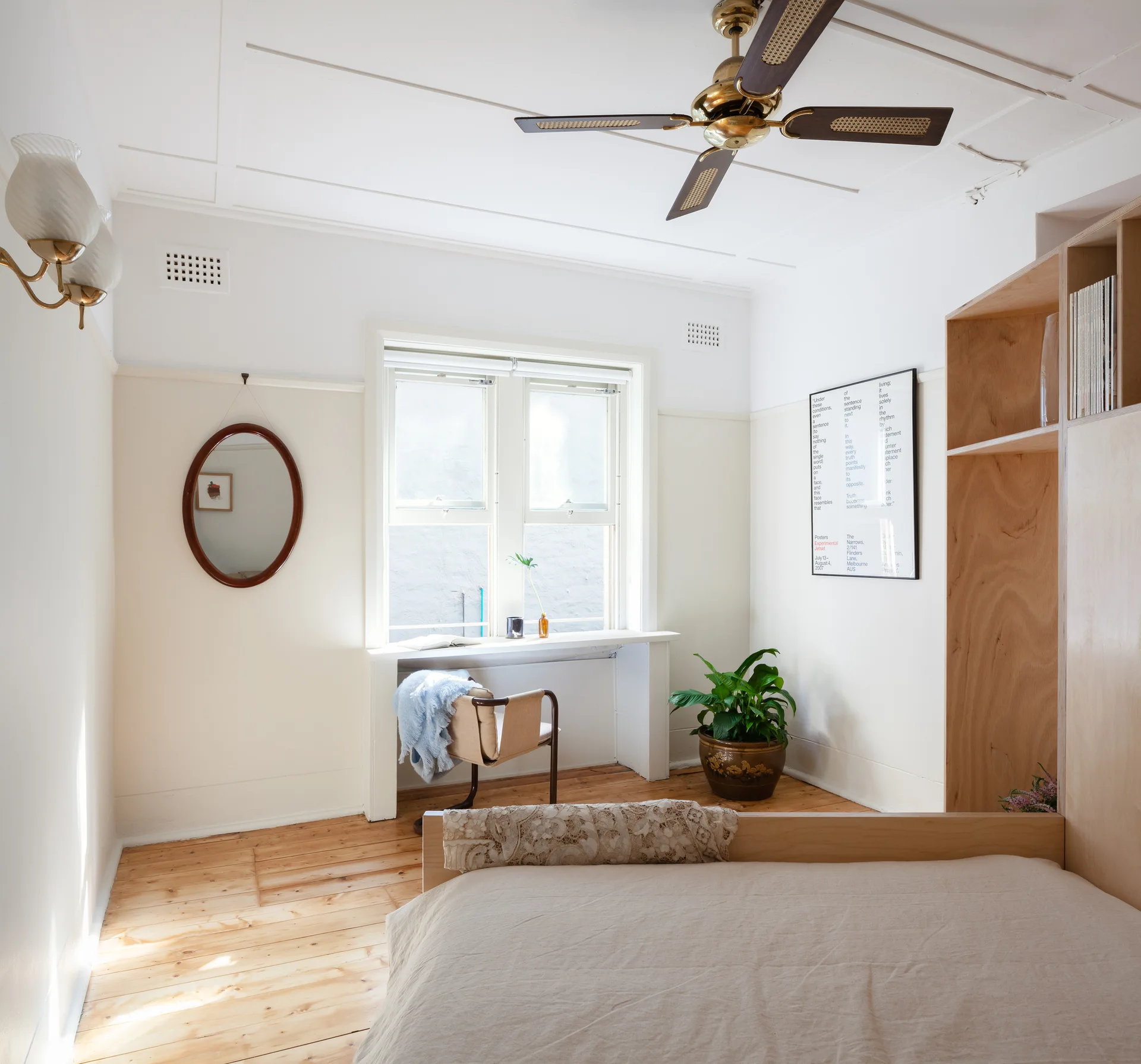 A minimalist bedroom with light pine floors, two-tone cream and white walls, a custom plywood storage unit, and a small desk by the window.